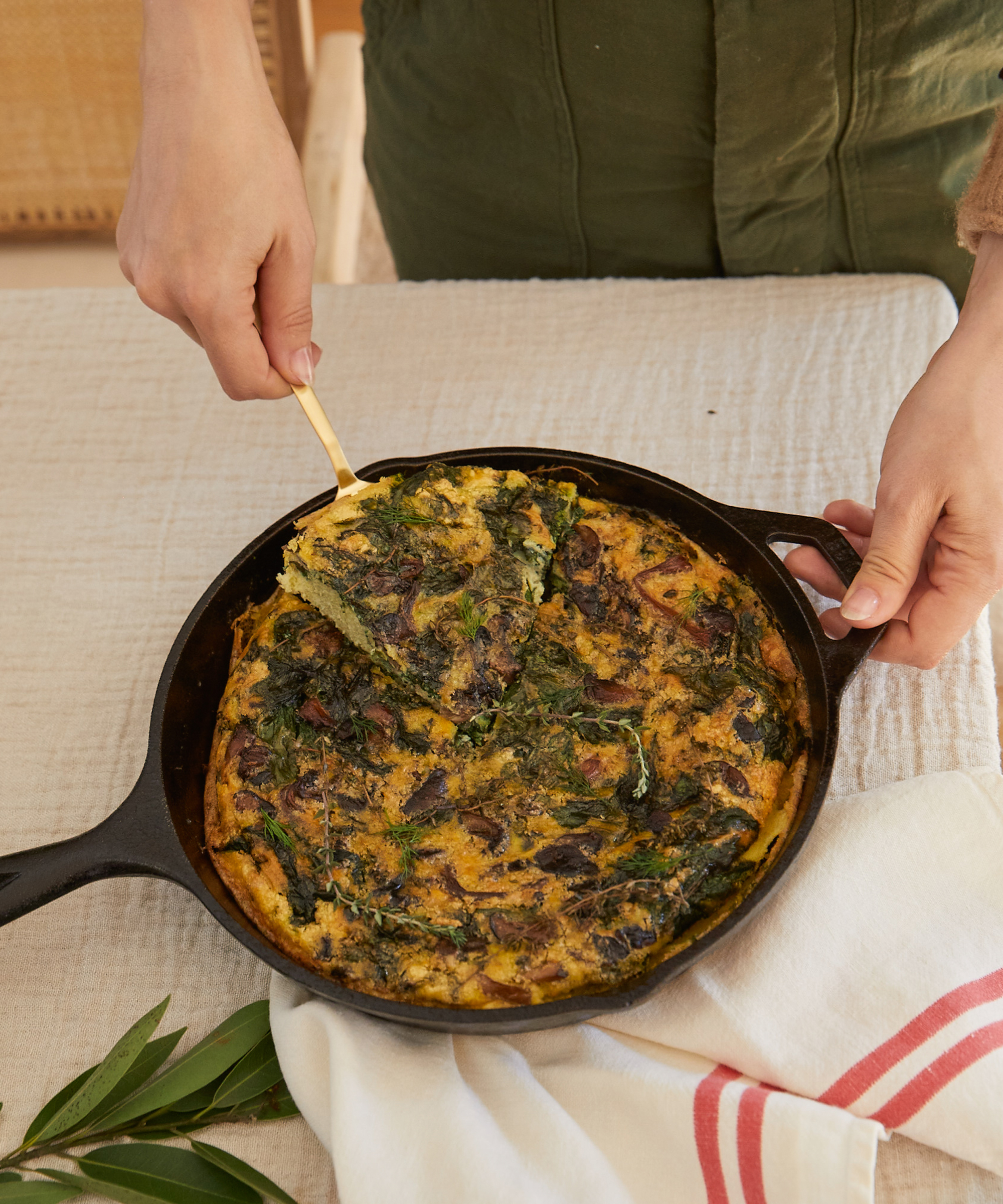 A person serves a slice of vegetable frittata, perfect for holiday recipes, from a black cast iron skillet using a gold utensil. The skillet rests on a beige tablecloth beside a red-striped white towel and fresh green herbs.