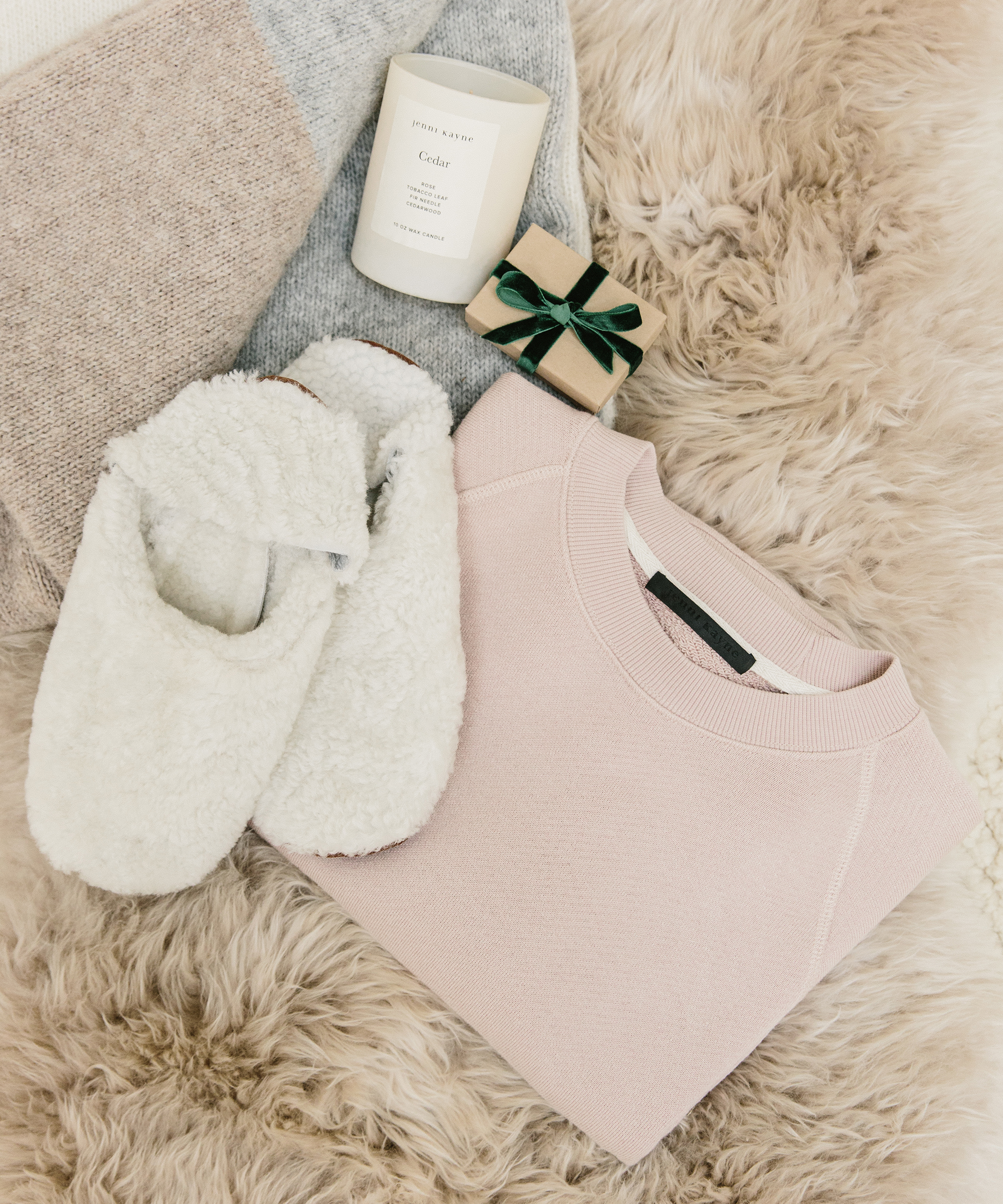 Jenni Kayne shearling slippers, blush sweatshirt, candle, blanket, and small wrapped box sitting on top of a sheepskin.