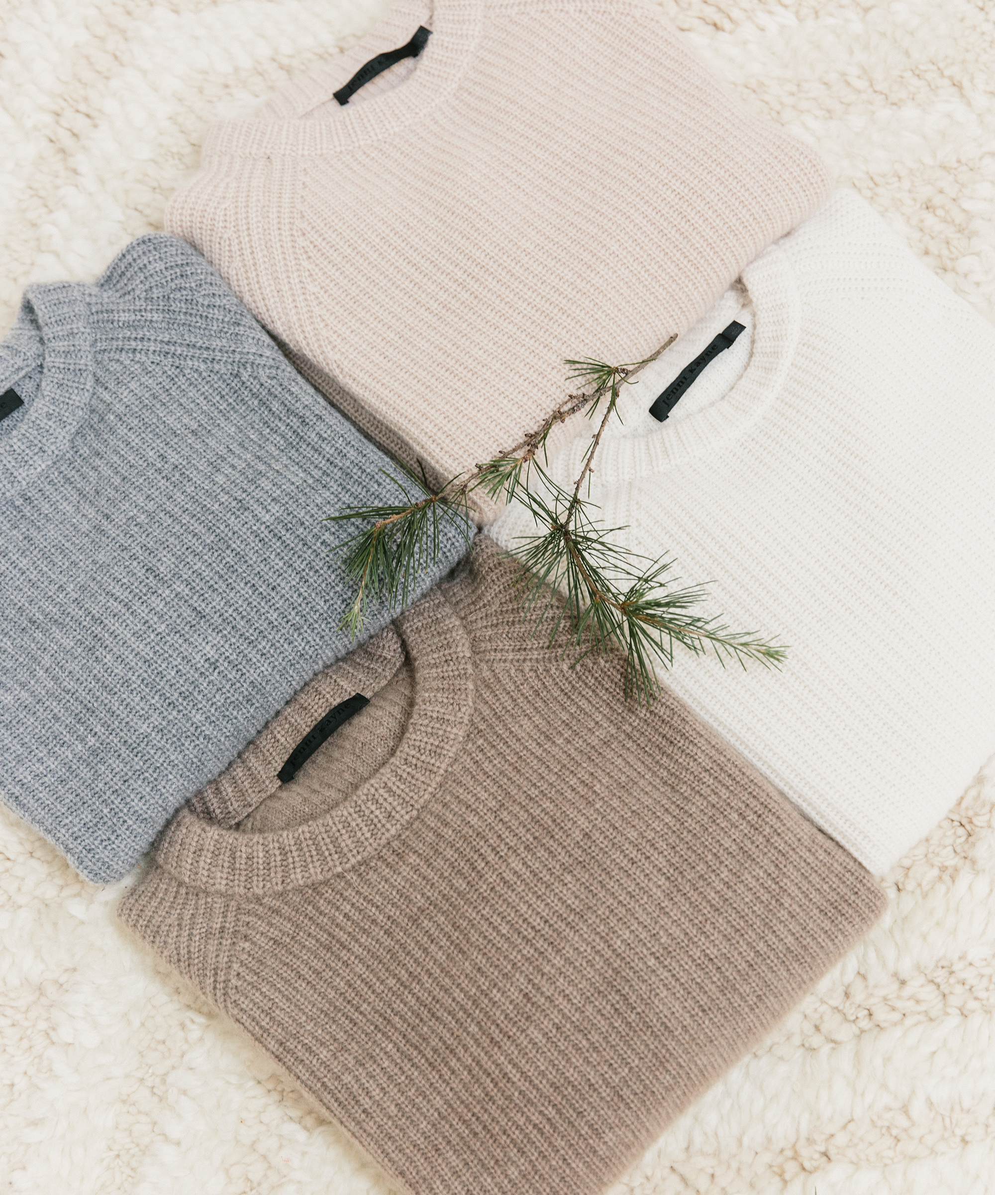 Four neatly folded knit sweaters in soft pastel colors—light pink, white, gray, and beige—are arranged on a textured cream surface with a small pine branch placed on top, making them perfect for last minute gifts.