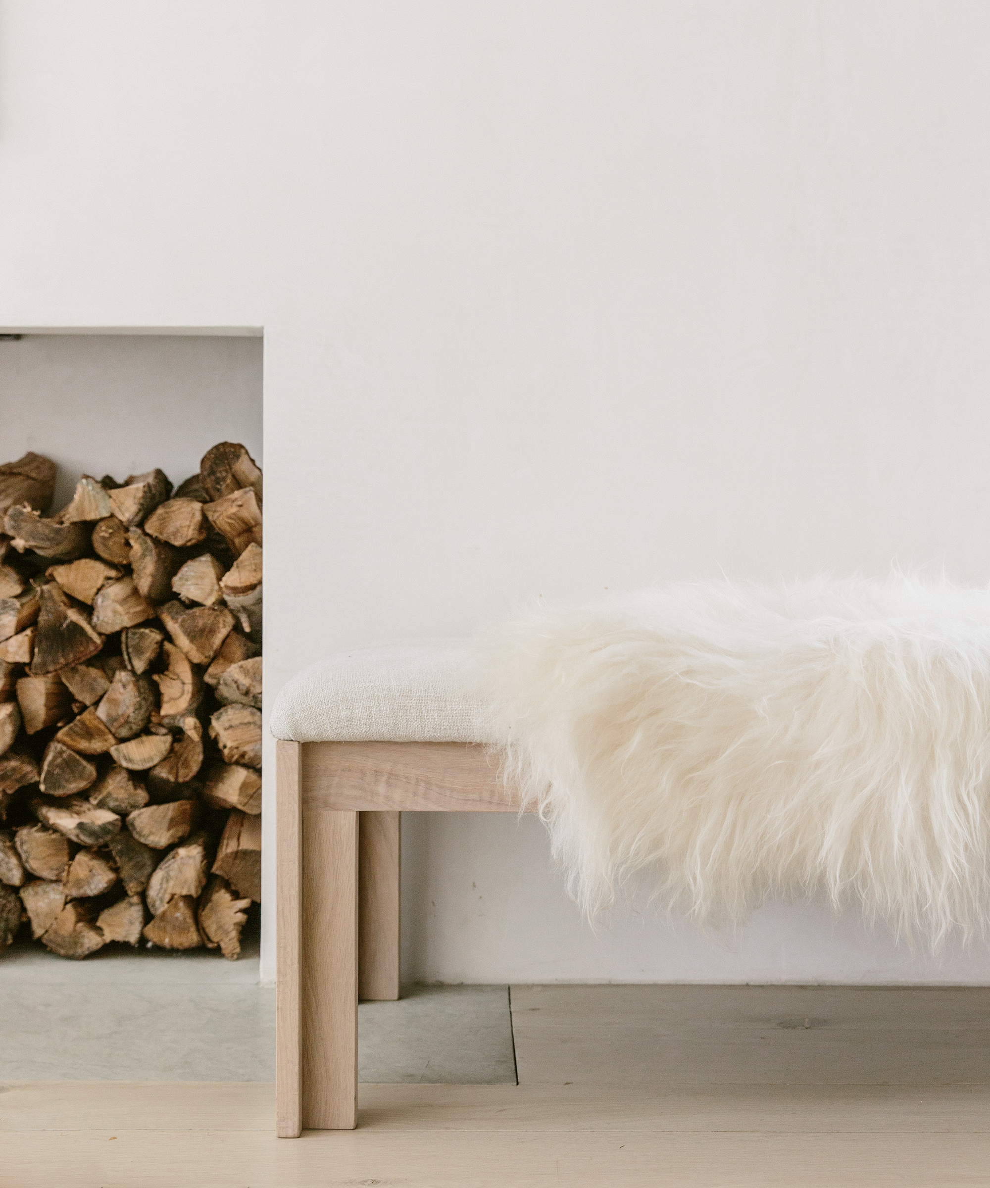 Living room shot of the boucle upholstered bench with a white sheepskin draped on top, sitting beside a stack of firewood.