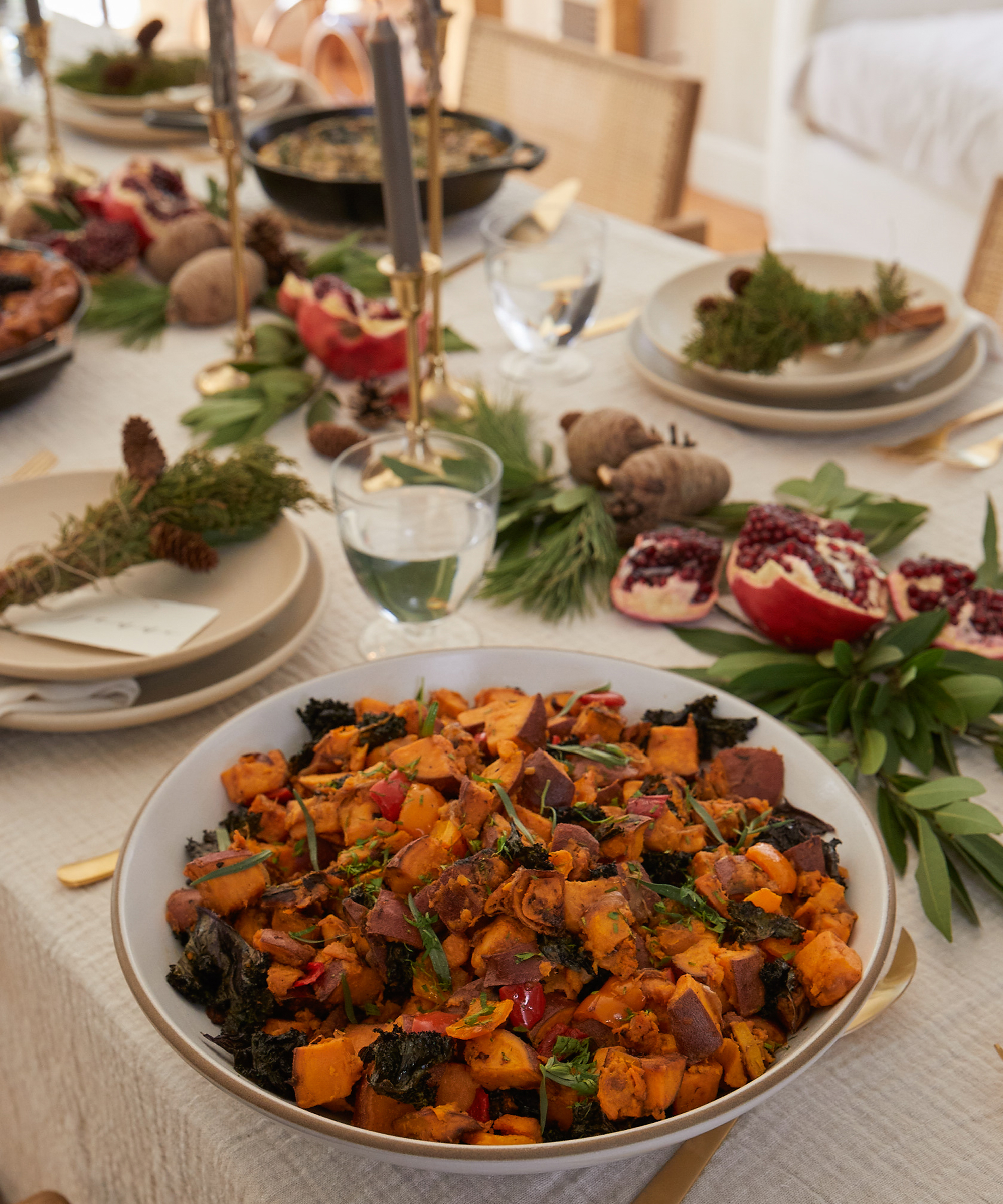 A festive dining table set with plates, glasses, and greenery showcases a large bowl of roasted vegetables in the foreground—perfect for holiday recipes—alongside pomegranate halves and decorative candles as a stunning centerpiece.