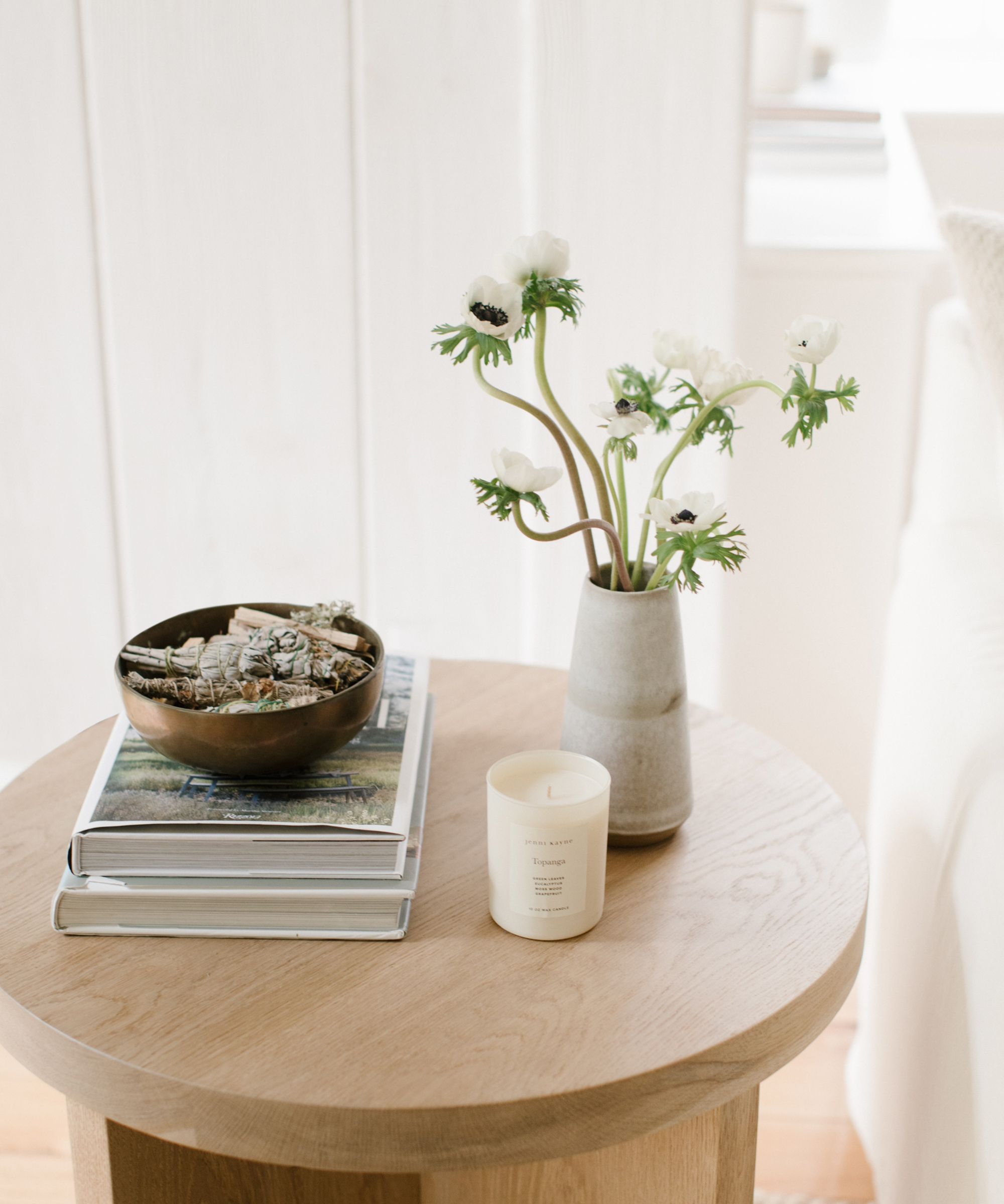 Small oak side table with a stack of two books, a ceramic bowl, a candle, and a vase with flowers.