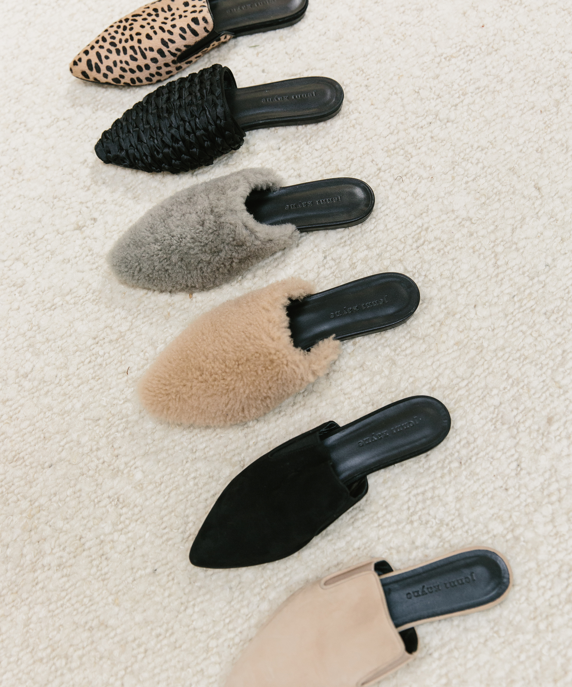 Six pairs of slip-on mules with different textures and colors—perfect for your new year style resolutions—feature animal print, woven black, gray fur, beige fur, black suede, and beige leather, all arranged in a line on a cream-colored carpet.