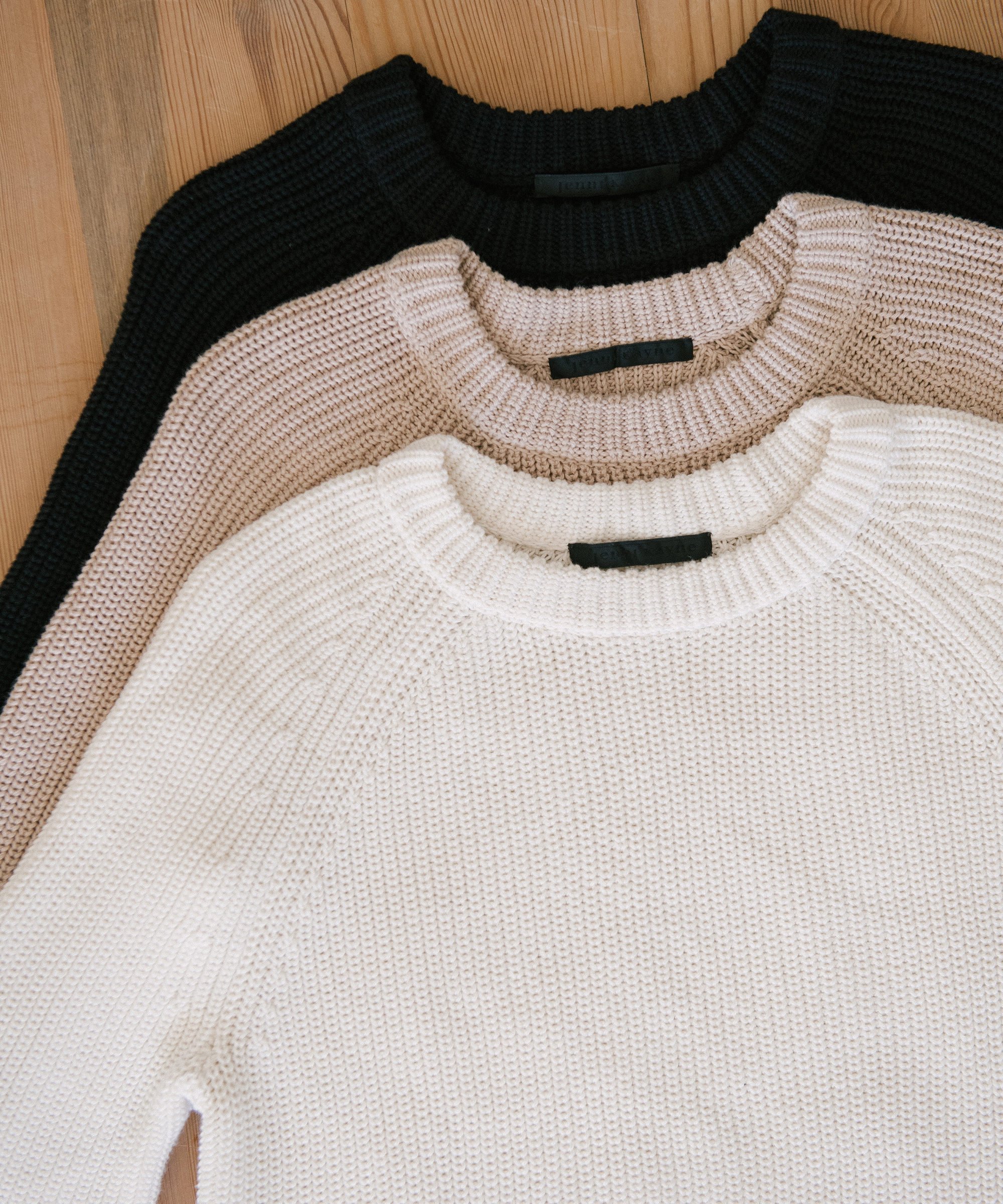 Jenni Kayne cotton fisherman sweaters in oatmeal, white, and black on a wooden table.