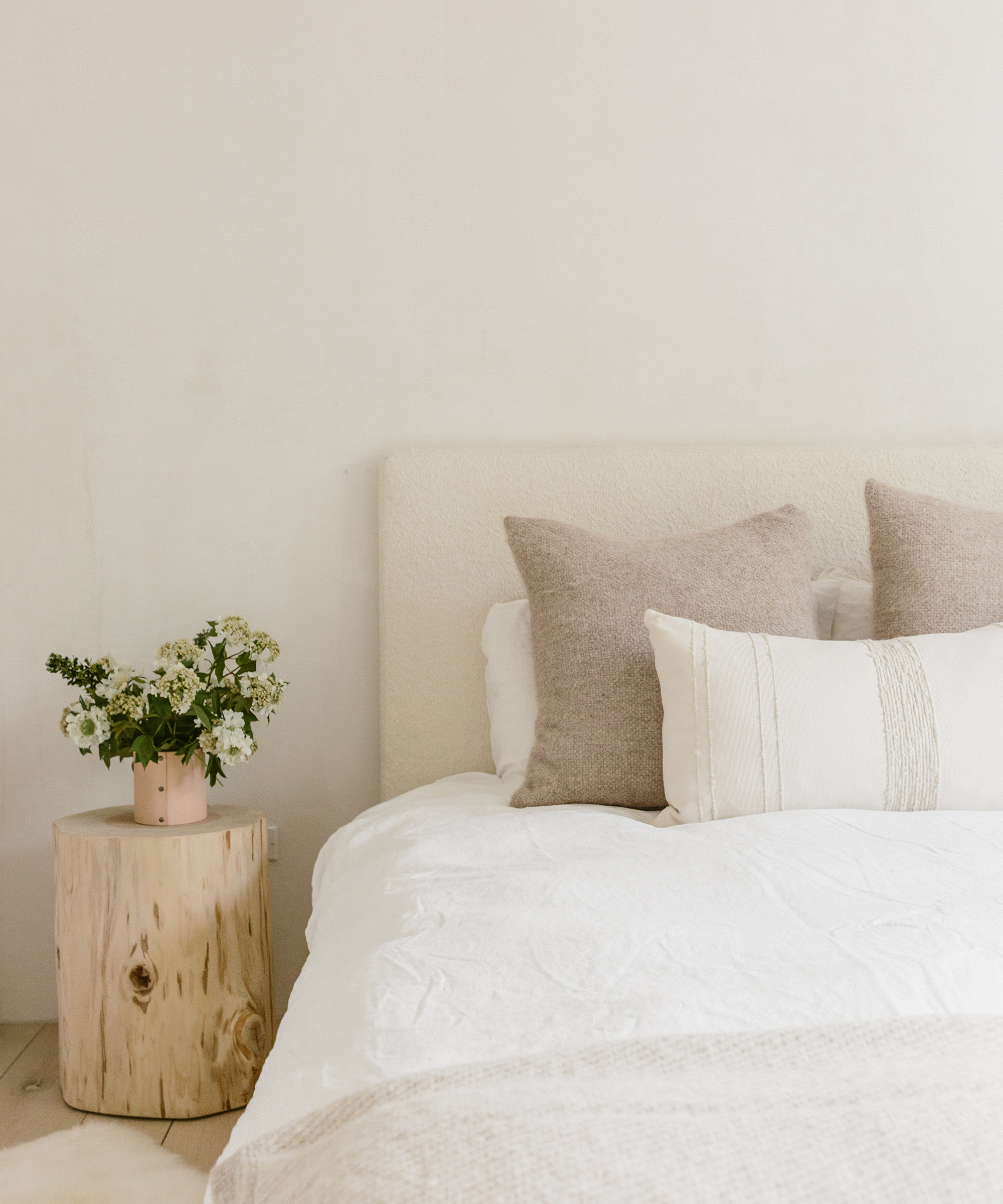 Neutral bedroom with Jenni Kayne cedar stump with a bud vase, flowers, and lamp on top sitting next to a Jenni Kayne Pacific Bed and white linens.