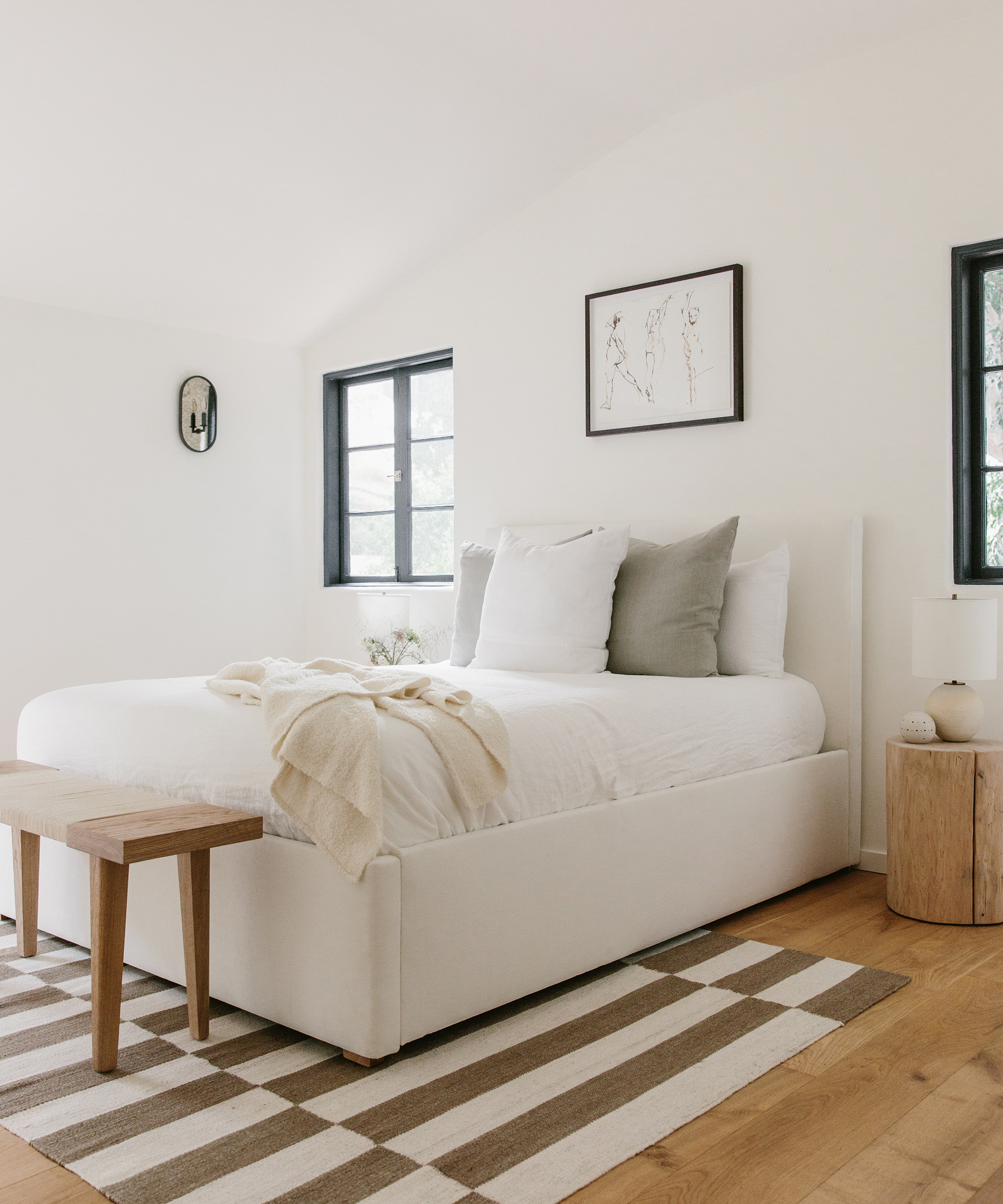 Bedroom with jenni kayne bungalow rug, ivpry pacific bed, white linens, assorted jenni kayne pillows, oak day bench at end of bed, and cedar stump as side table.