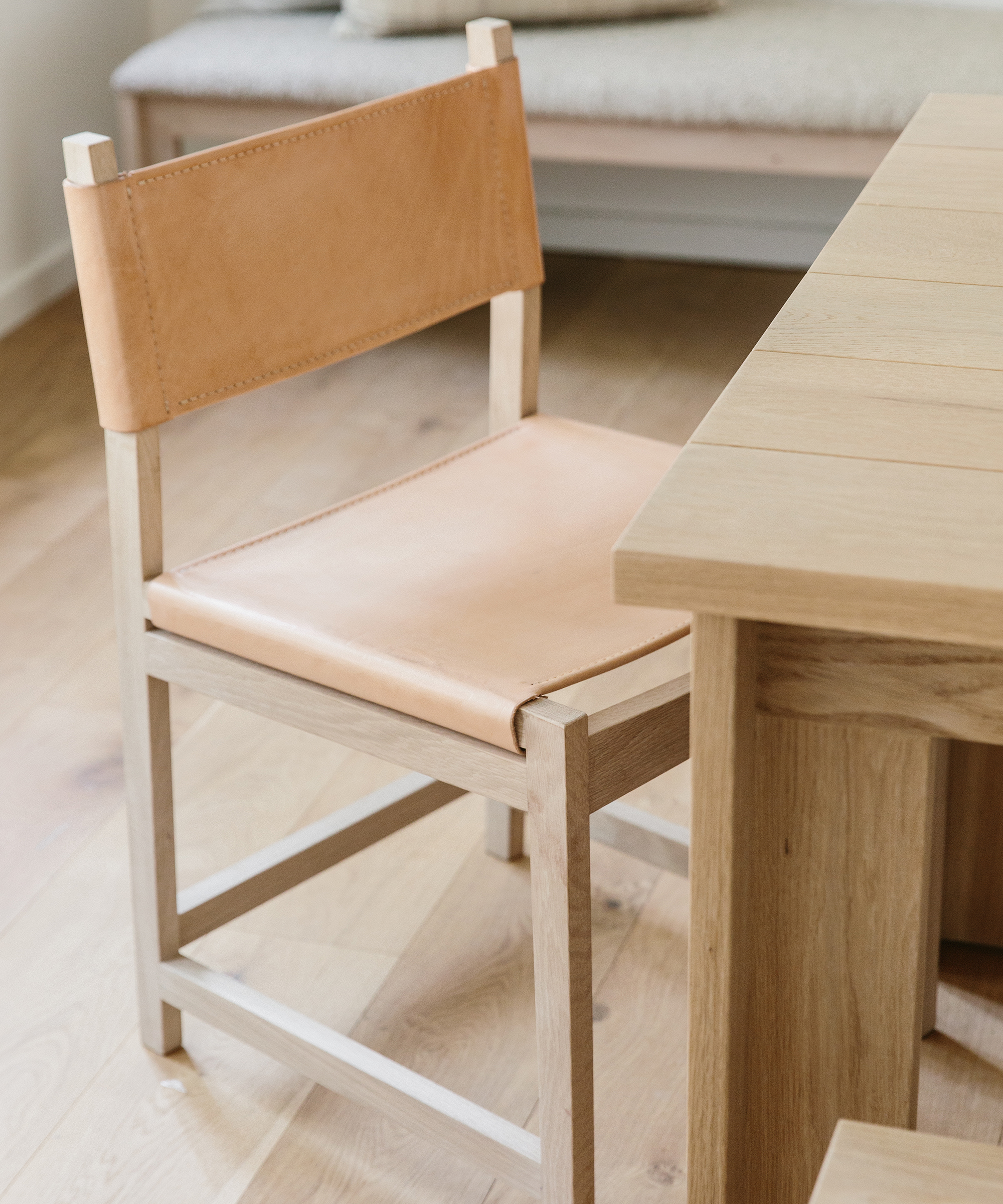 A light wooden chair with tan leather seat and backrest, part of stylish dining room furniture, is positioned next to a matching wooden table on a light wood floor. A cushioned bench is visible in the background.