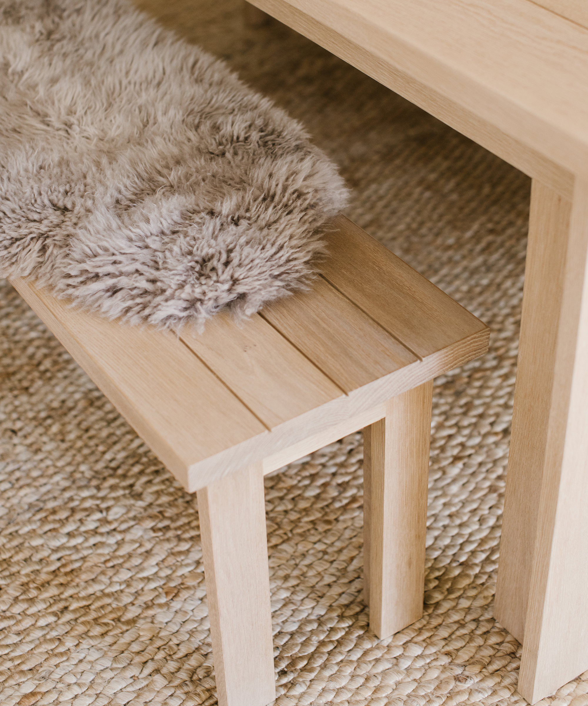 A light wooden bench, part of elegant dining room furniture, with a soft grayish-brown faux fur throw sits on a textured woven rug next to a matching wooden table.