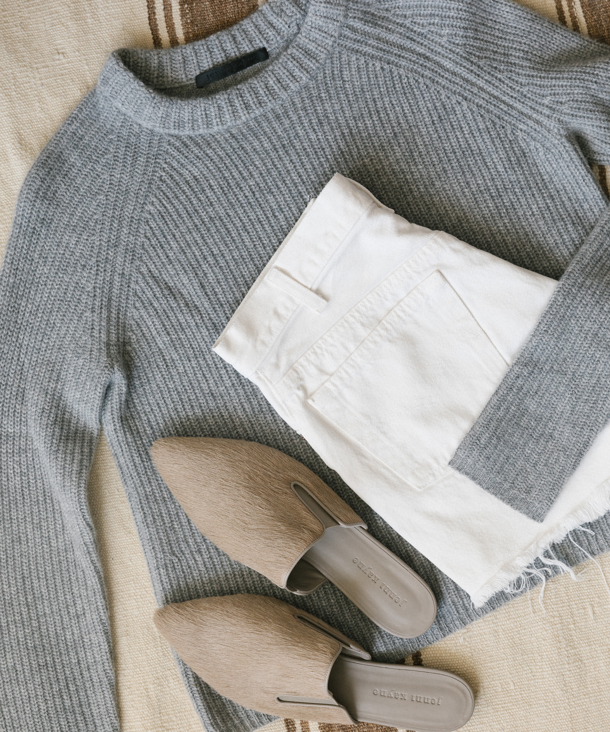 A neatly folded light gray all-season sweater, white jeans, and a pair of taupe-colored, pointed-toe mules are arranged on a beige textured surface.