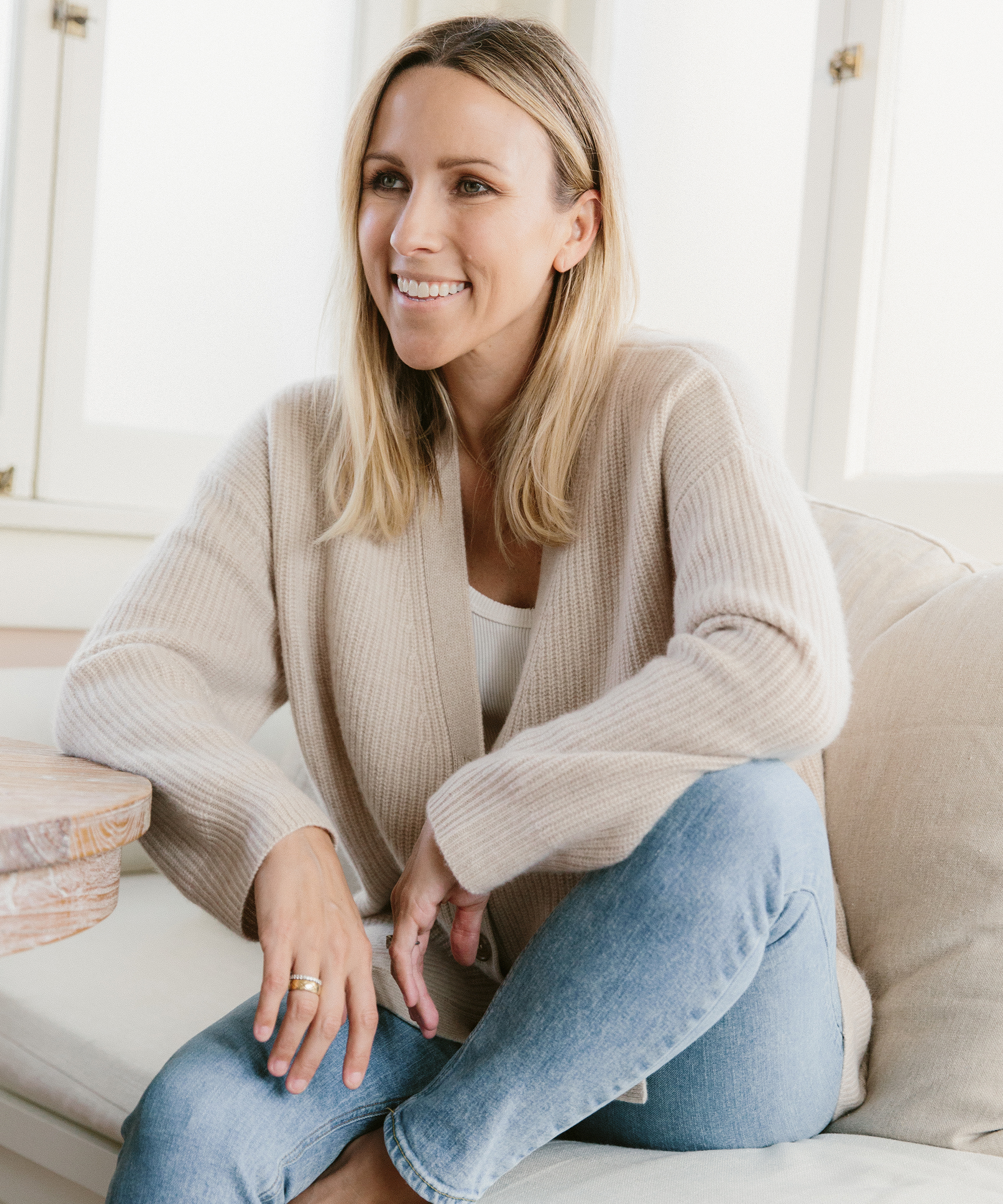 Jacey Duprie sitting in her living room smiling while wearing a jenni kayne cashmere cocoon in oatmeal.