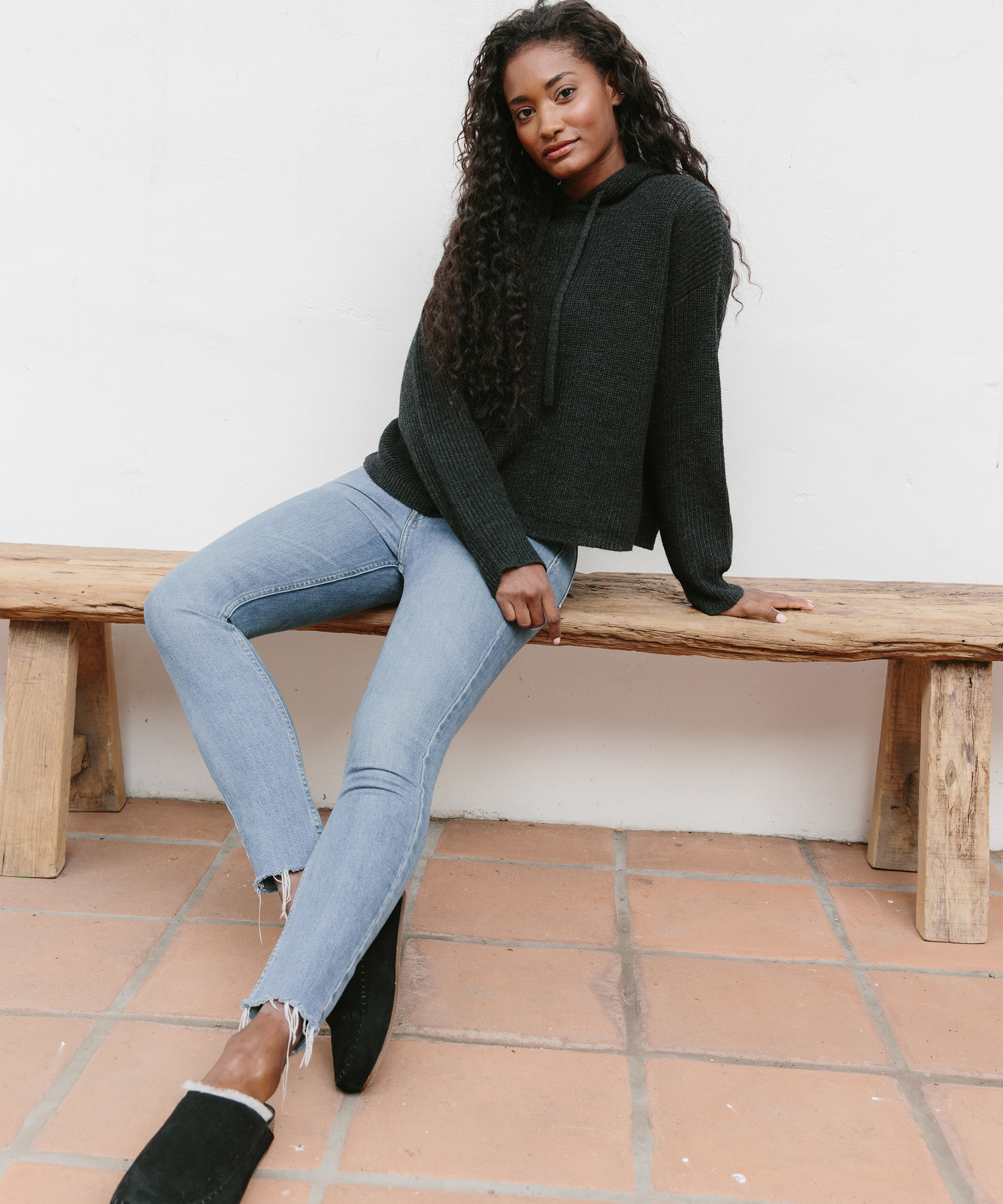 A woman with long curly hair sits on a wooden bench against a white wall, showcasing one of her stylish work from home outfits: a dark sweater, light blue jeans, and black shoes. She crosses one leg over the other and looks relaxed.