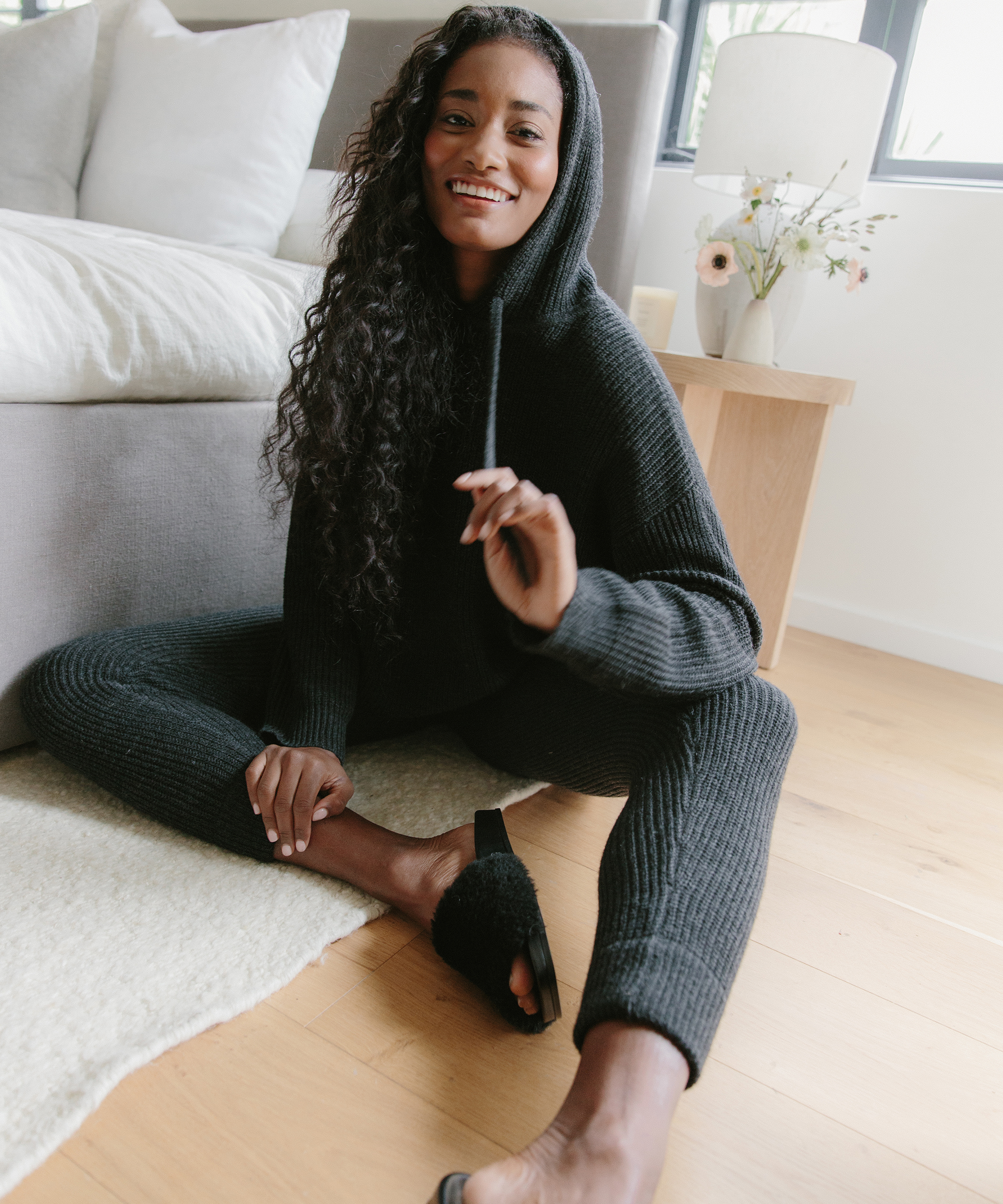 A woman with long curly hair sits on a wooden floor, smiling and wearing a dark, ribbed hooded sweater and matching pants—perfect work from home outfits. She is barefoot, with one slipper on, in a cozy, bright living room.