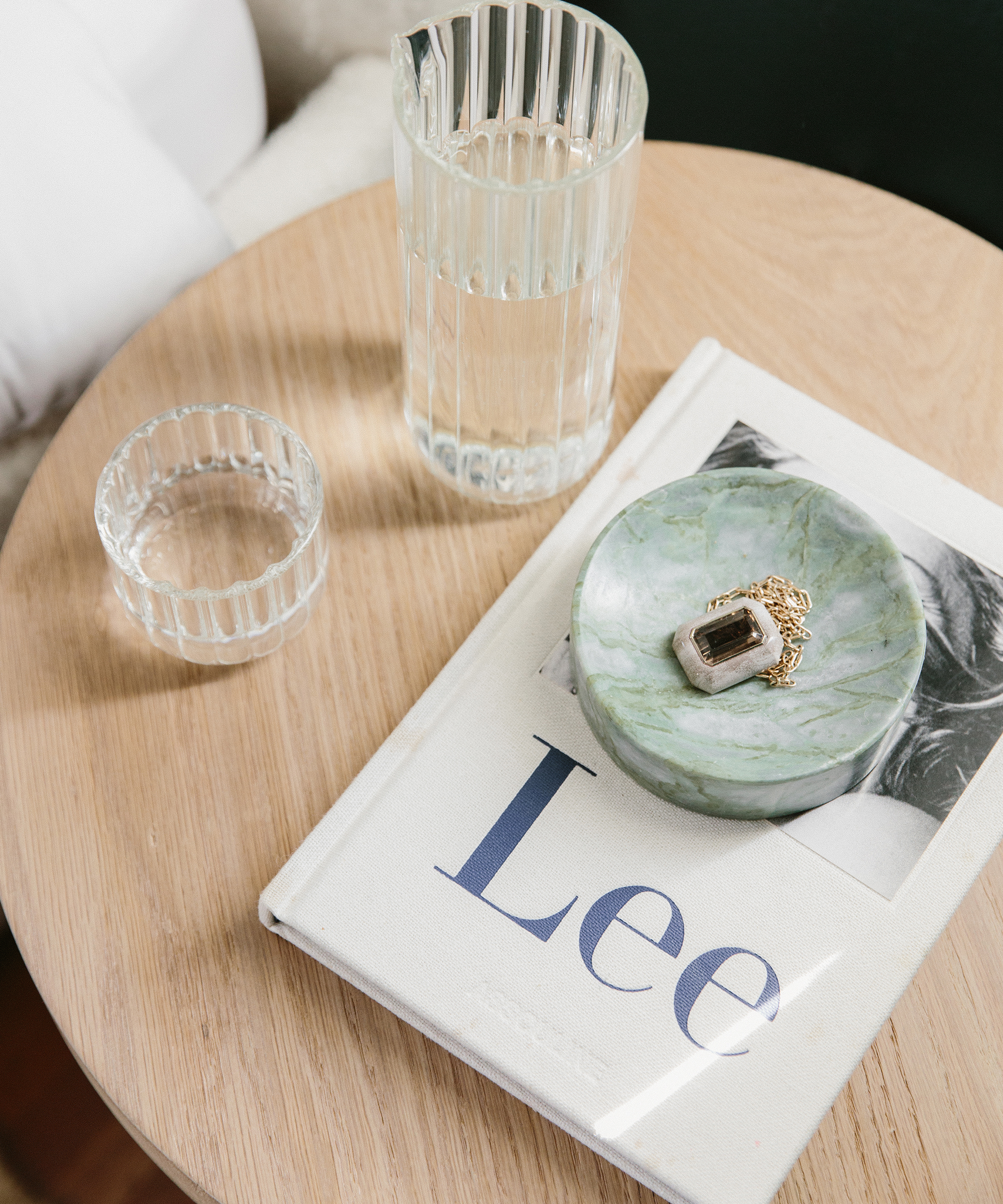 Close up shot of small oak side table with glass of water, coffee table book, and ceramic bowl with a necklace inside.