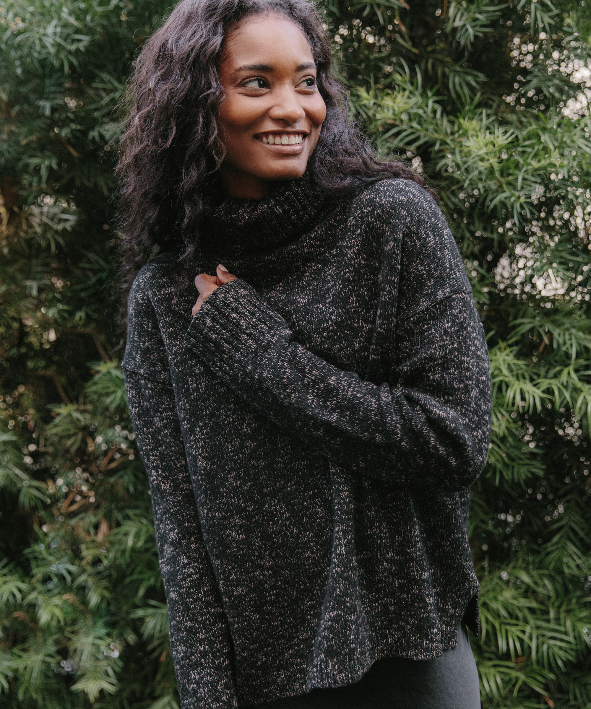A woman with long curly hair smiles while standing outdoors in front of green foliage, wearing a dark, cozy turtleneck sweater—one of her favorite work from home outfits.
