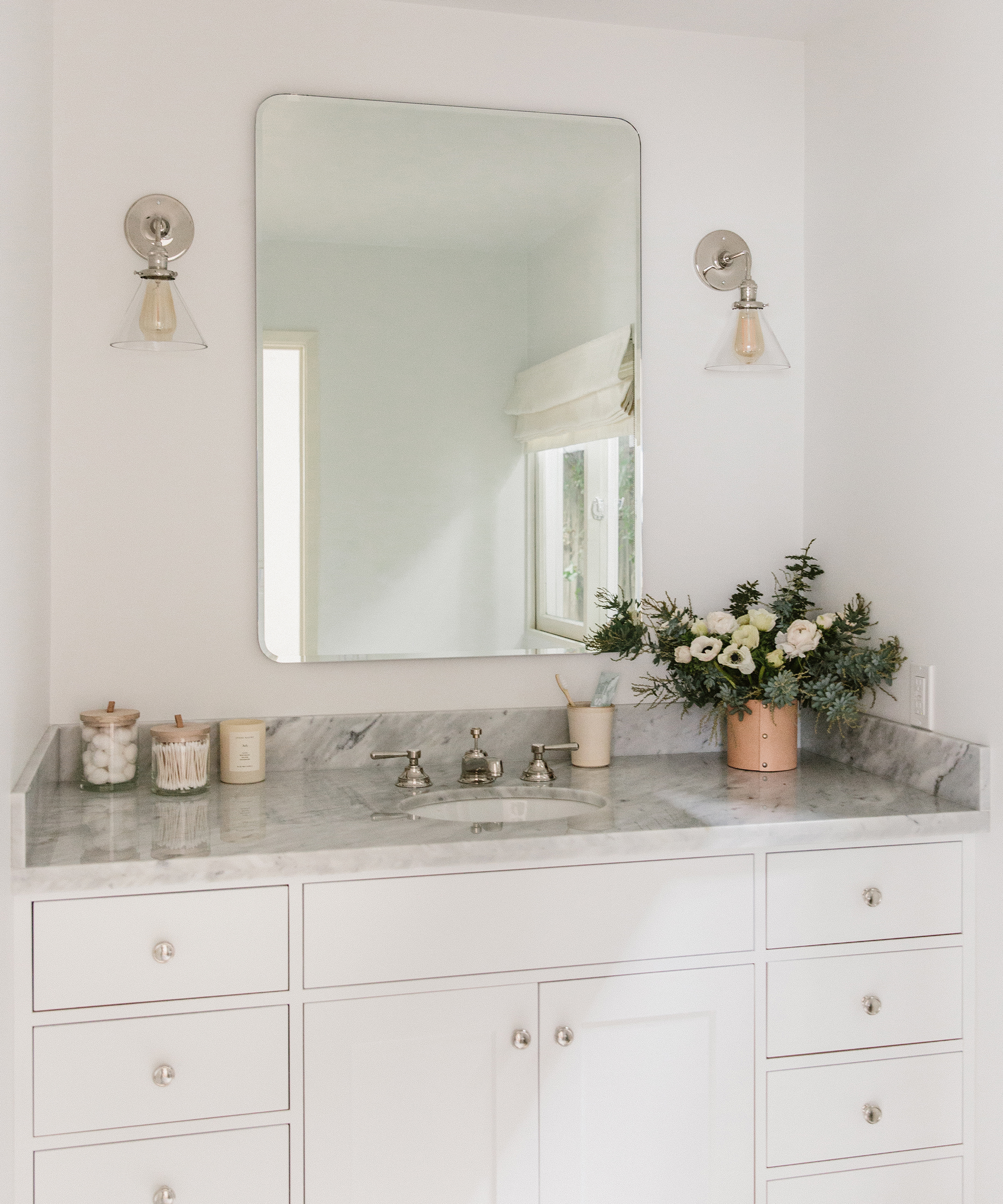A bright, minimalist bathroom remodel features white cabinets, a marble countertop, silver fixtures, a rectangular mirror, two wall sconces, and a vase of white flowers next to candles and jars.