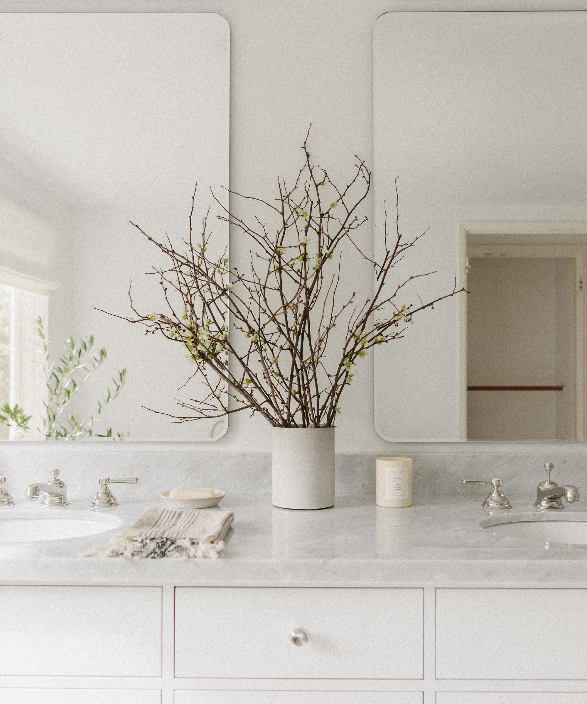 A minimalist bathroom counter with a white marble top, two sinks, a vase of bare branches, a candle, a soap dish, and two mirrors reflecting the neutral, light-filled space—perfect inspiration for your next bathroom remodel.