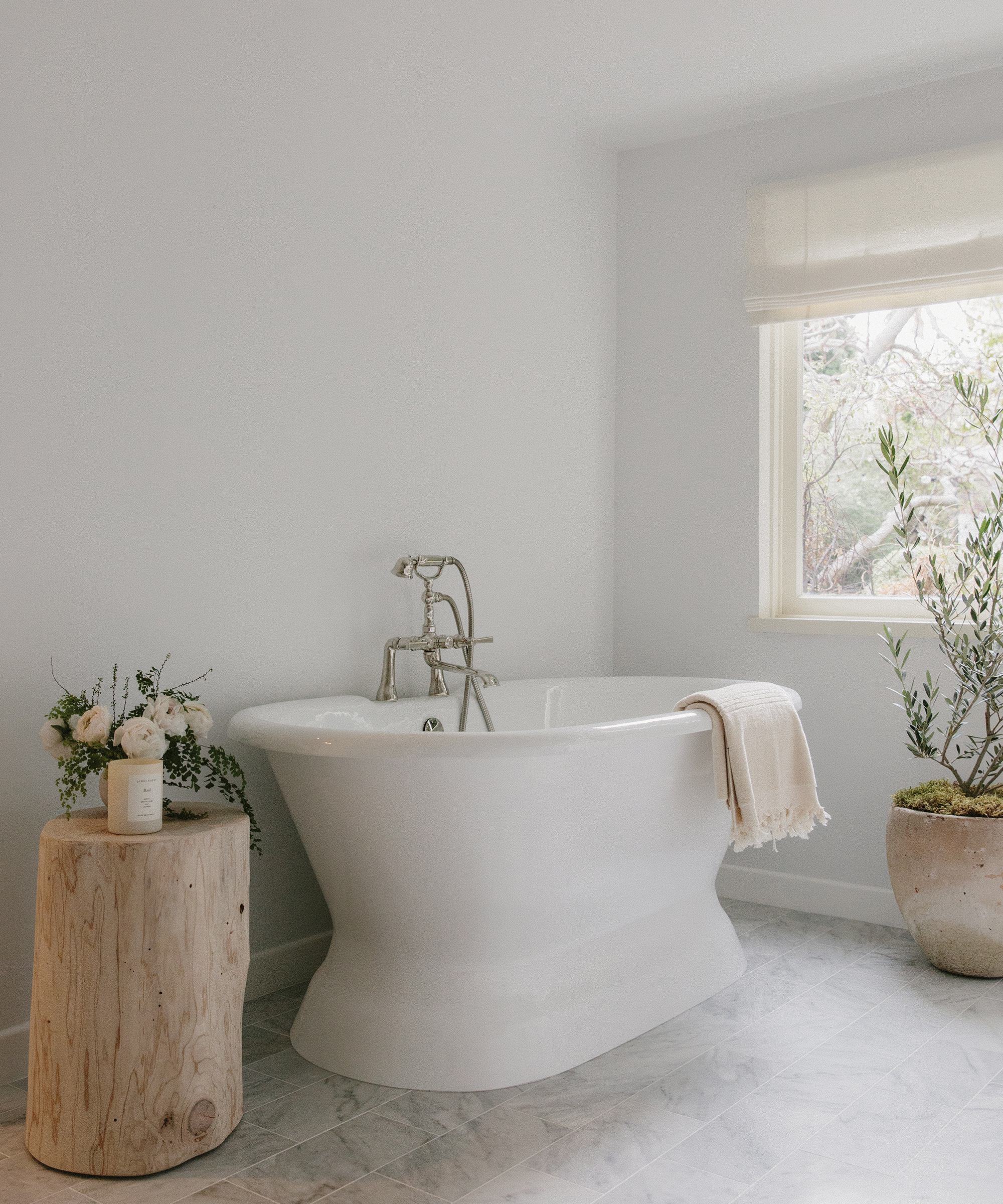 A modern bathroom remodel with a white freestanding bathtub, a beige towel draped over the side, a wooden stump table holding flowers and a candle, a large potted plant, and soft natural light from a window.