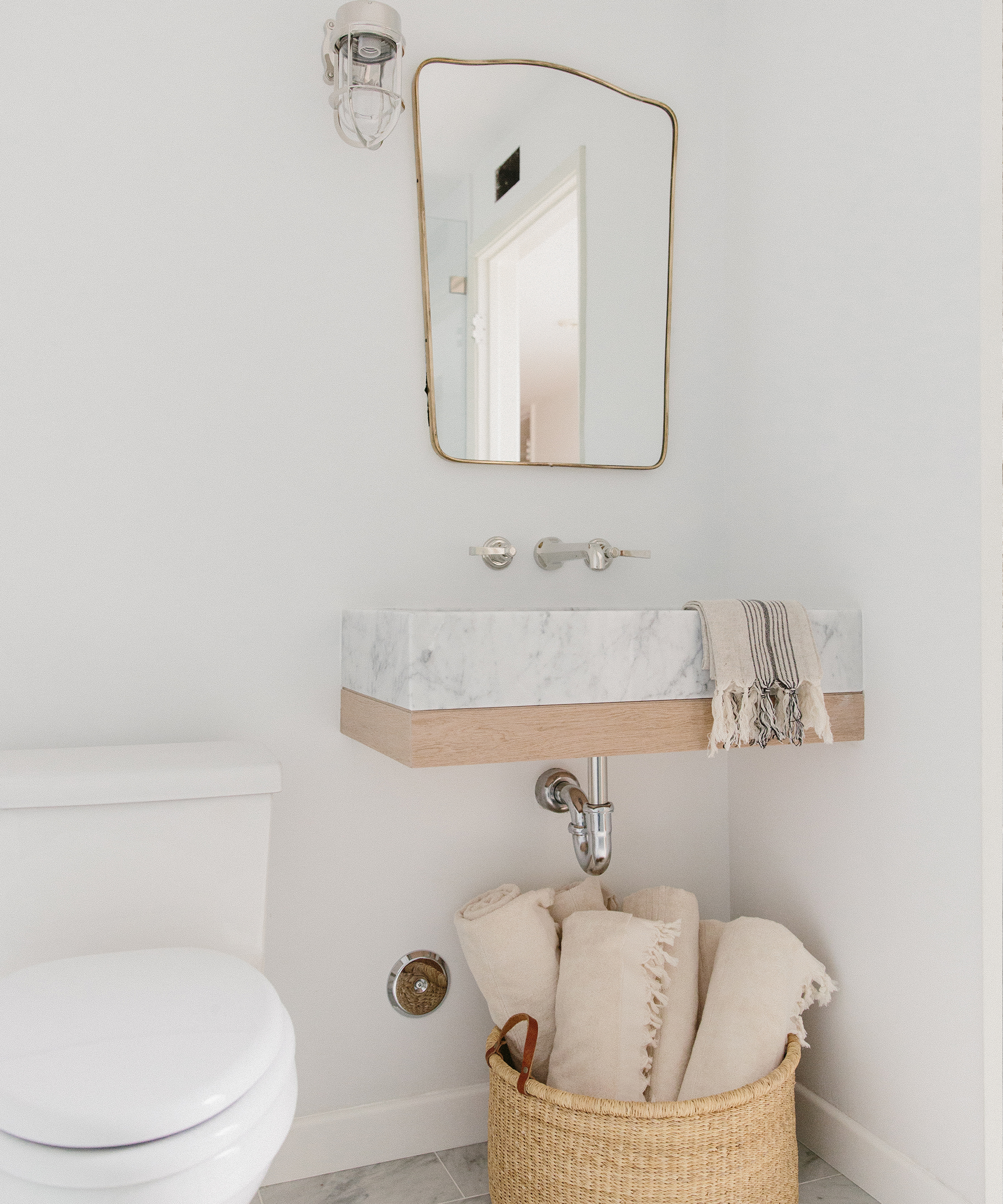 A minimalist bathroom remodel features a wall-mounted marble sink, a rectangular mirror, white walls, a toilet, and a woven basket below the sink holding rolled-up towels with a striped hand towel draped on the sink.
