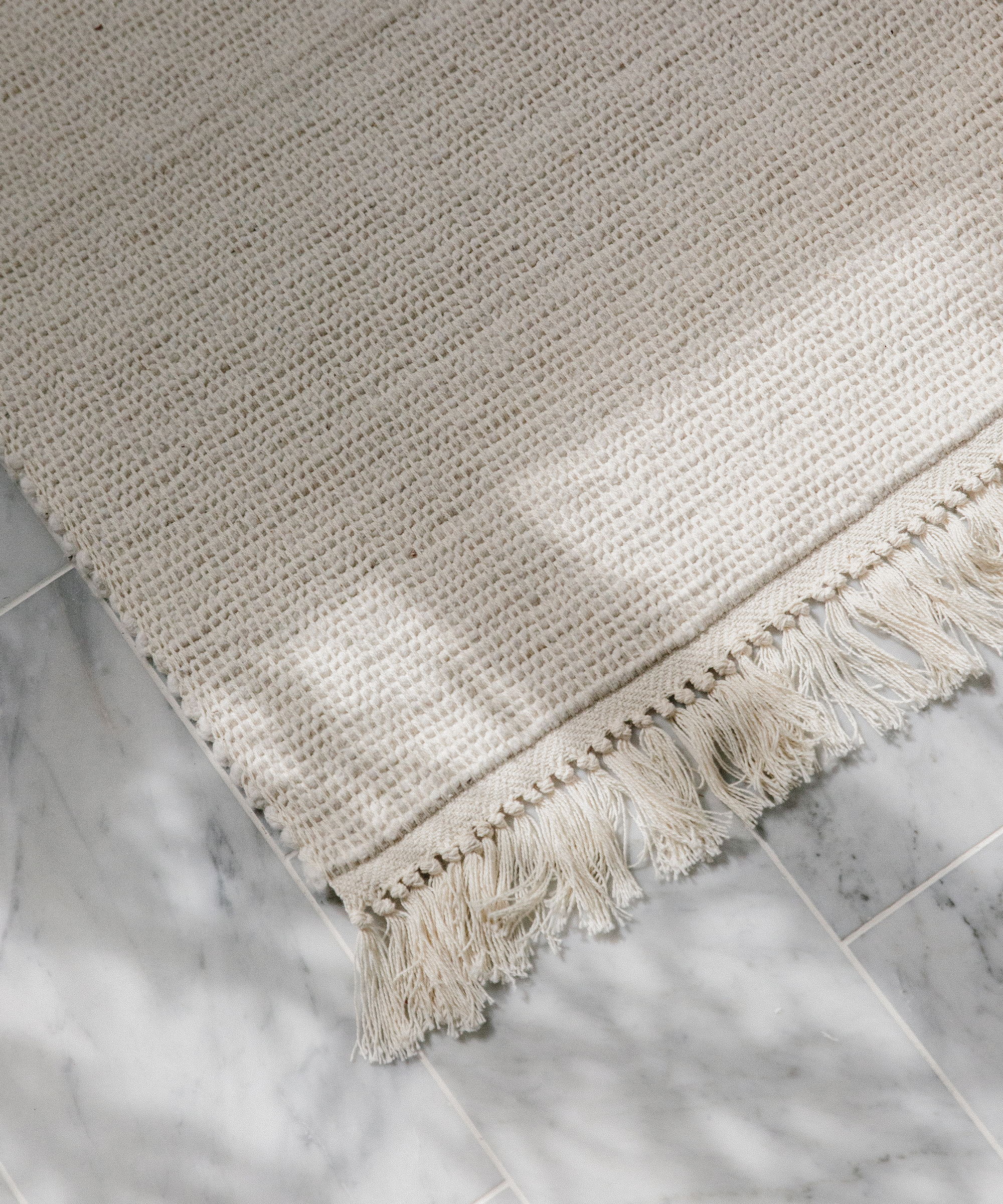 A cream-colored woven rug with fringe edges lies on a light gray marble tile floor, the perfect finishing touch for your bathroom remodel as sunlight casts soft shadows across the surface.