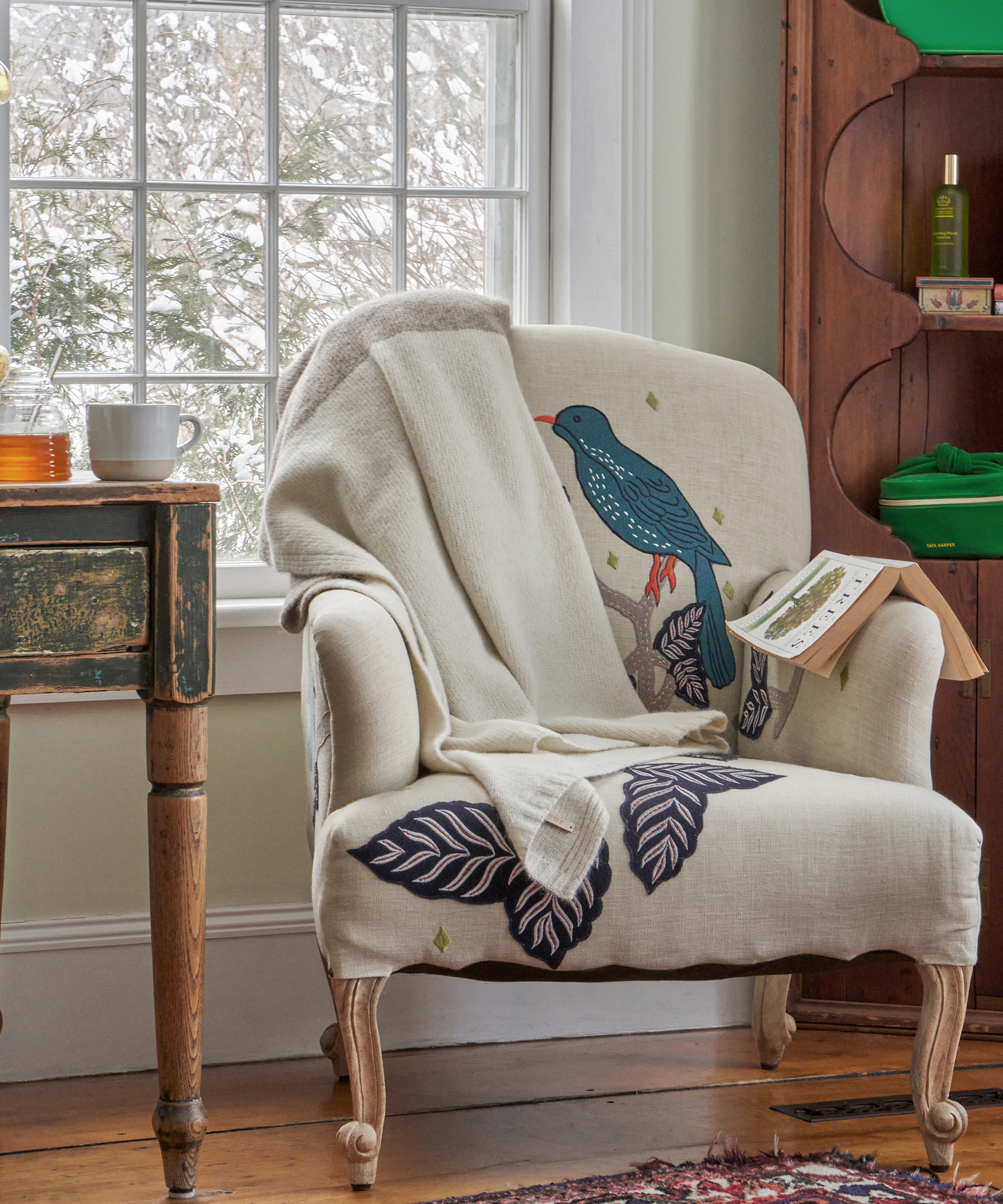 A cozy armchair with a bird and leaf design sits by a window with snowy trees outside. A cream-colored blanket, an open book, and a table topped with a cup, honey jar, and tata harper skincare complete the inviting scene.