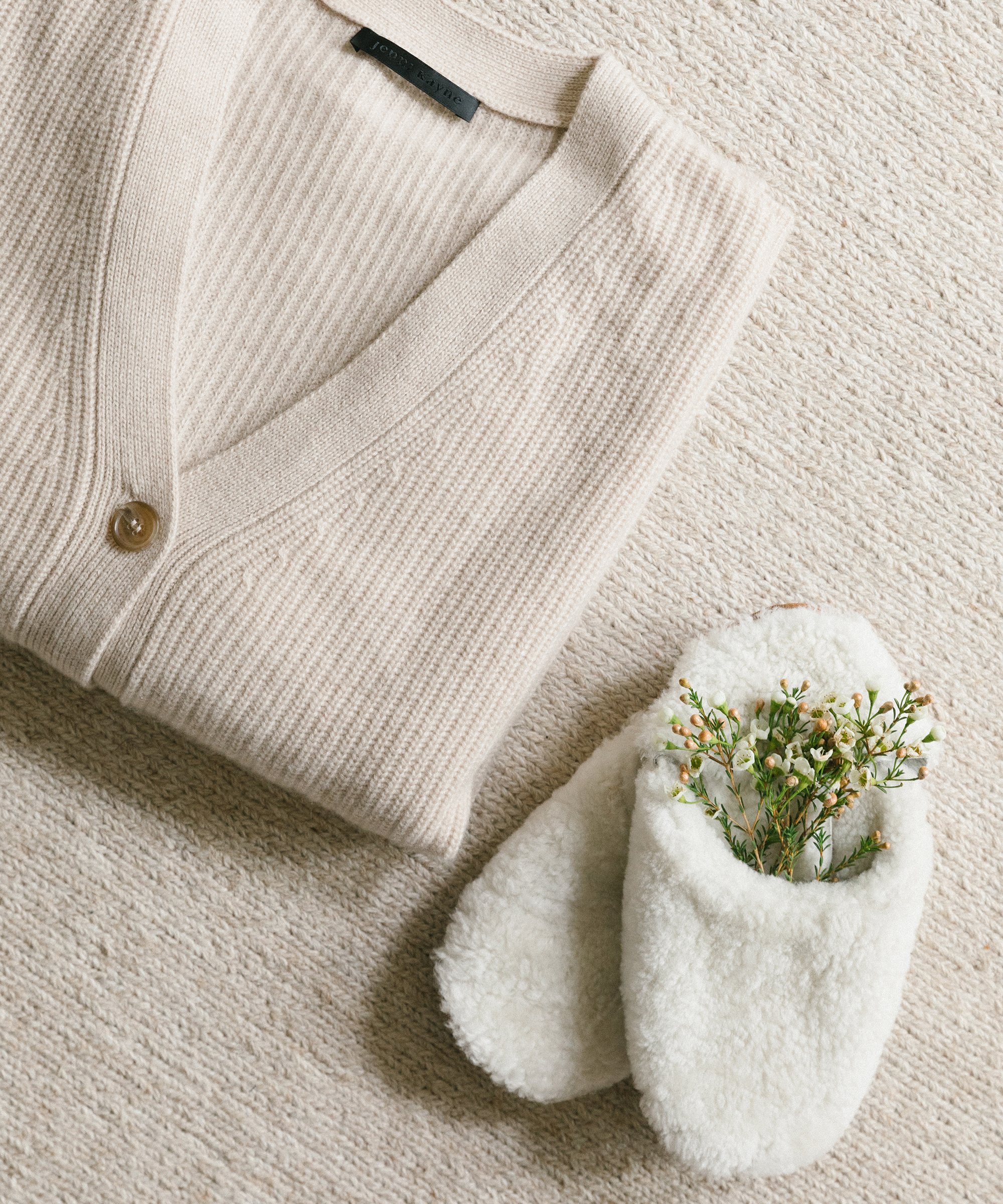 Jenni kayne cashmere cocoon sweater folded up next to shearling slippers with flowers sticking out of the sole.