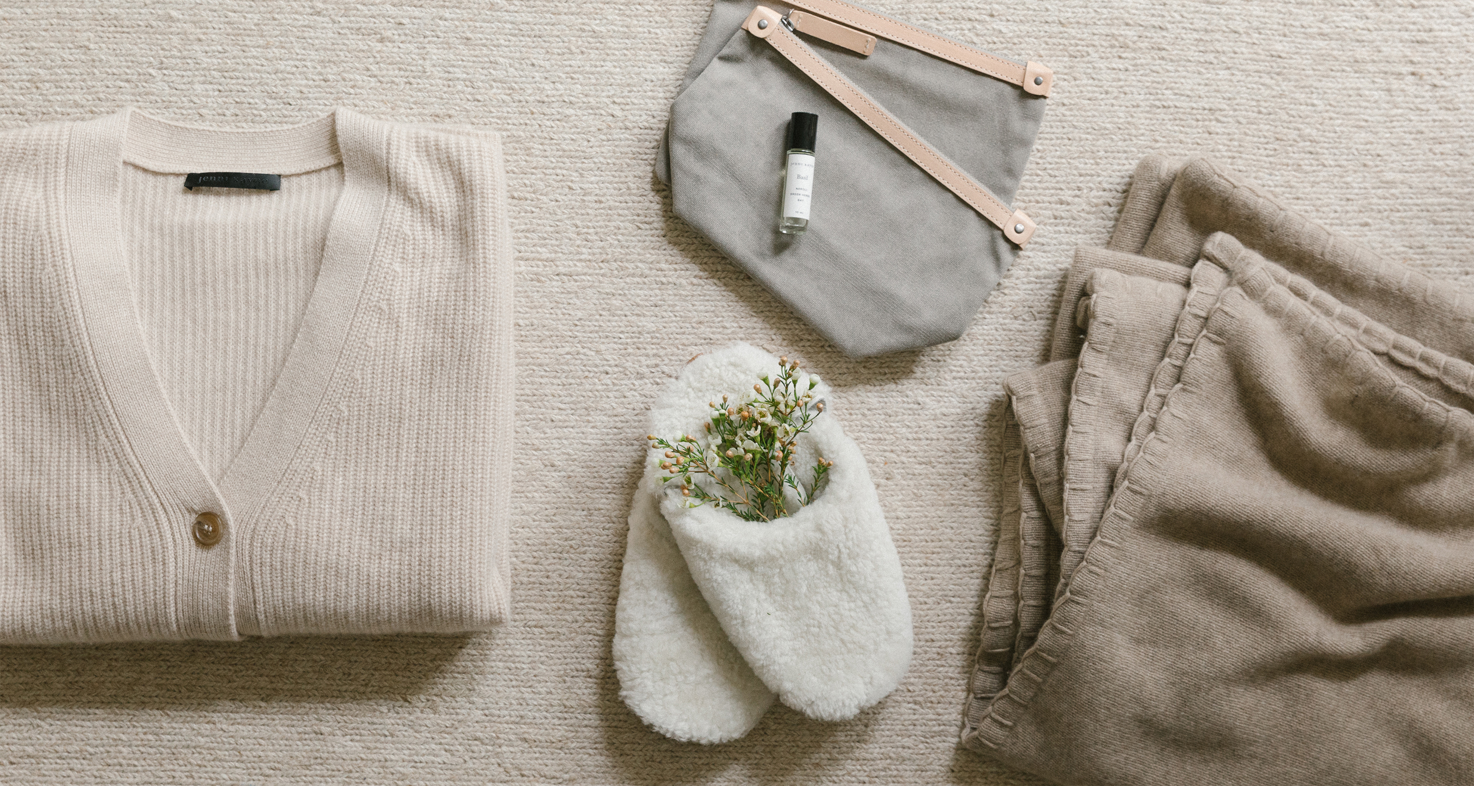 Laydown shot of jenni kayne cashmere cocoon sweater in oatmeal folded up next to jenni kayne shearling slippers, cotton travel pouches, roll-on, and tahoe cashmere blanket.