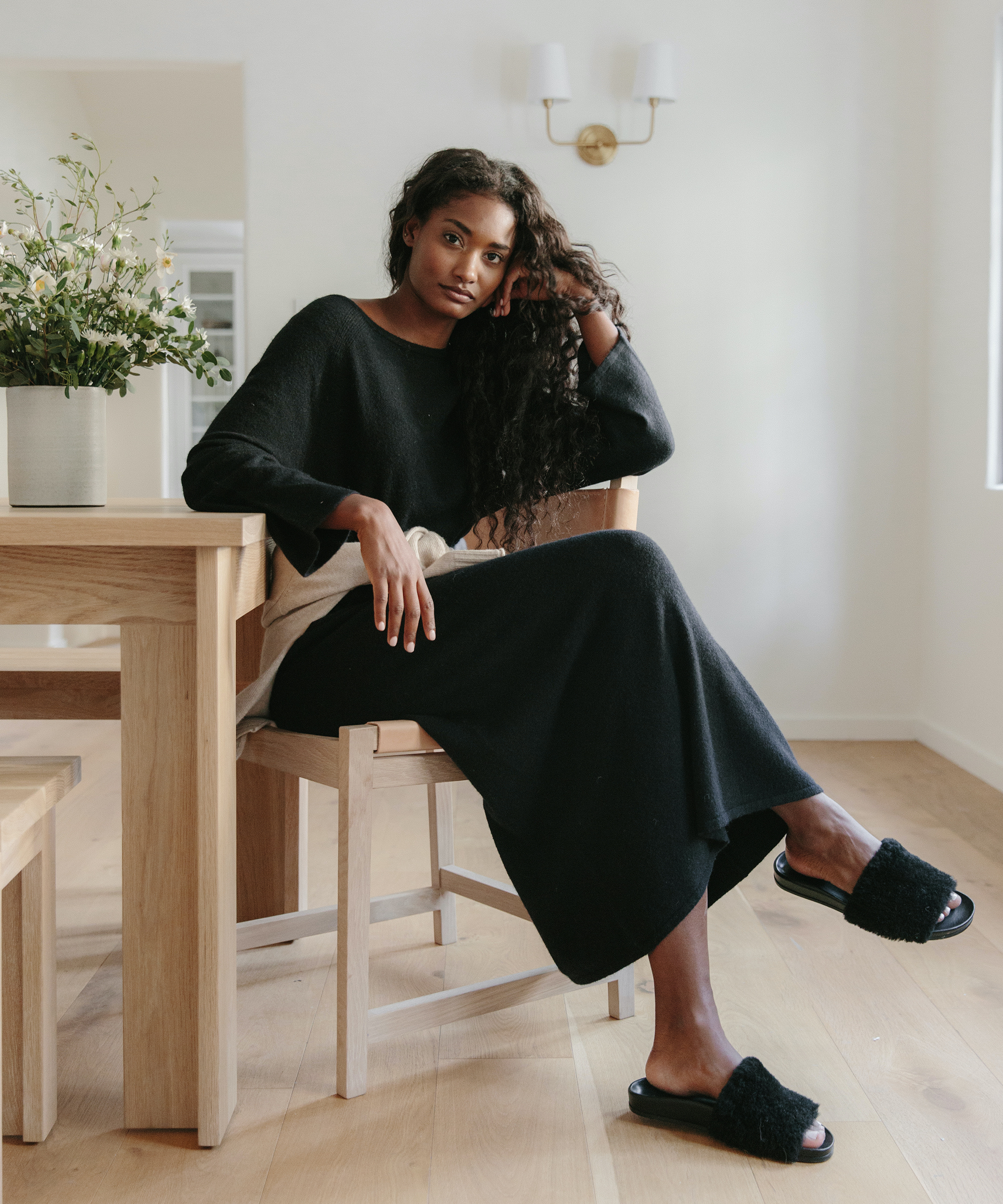 A woman with long curly hair sits at a wooden dining table in a bright room, wearing luxury loungewear—a black long-sleeve dress and fuzzy slippers. She rests her head on one hand and looks calmly toward the camera.