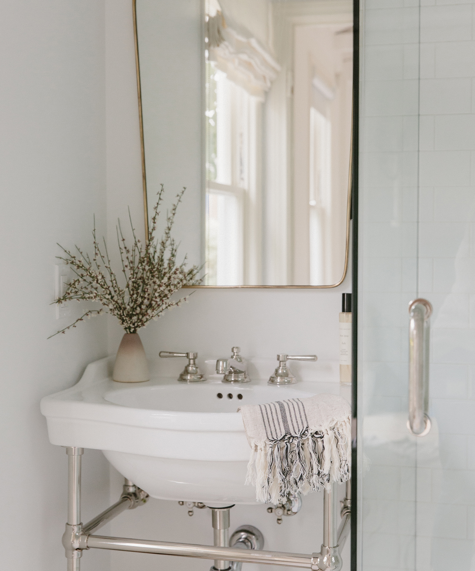 Small white bathroom sink with silver fixtures, a striped hand towel draped over the basin, a beige vase with branches, and a rectangular mirror above the sink—perfect touches for your bathroom remodel. A glass shower door is partially visible to the right.