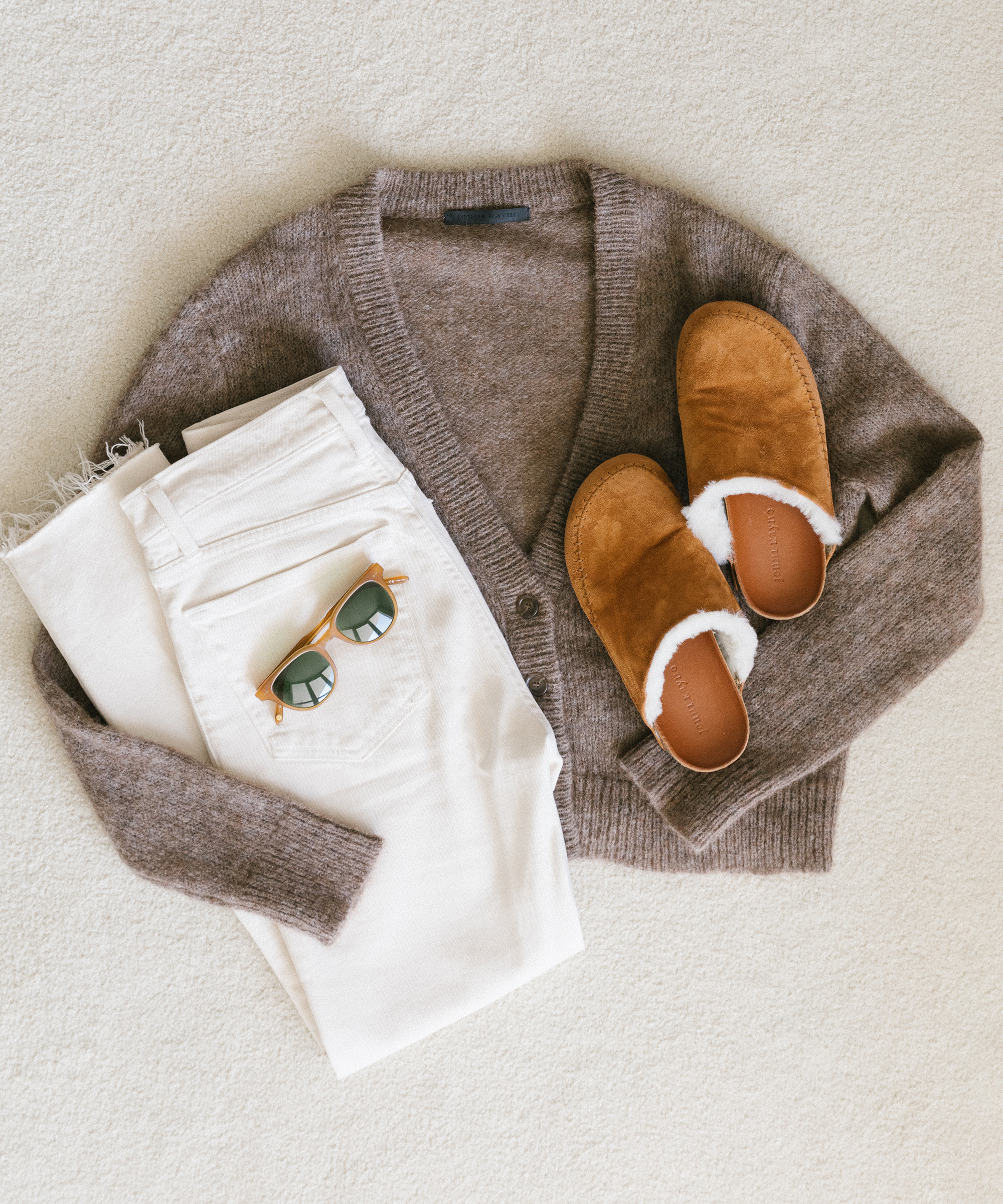 A cozy flat lay featuring a brown knit cardigan, white jeans, brown shearling slippers, and round sunglasses arranged neatly on a light surface—perfect for spring outfit ideas.