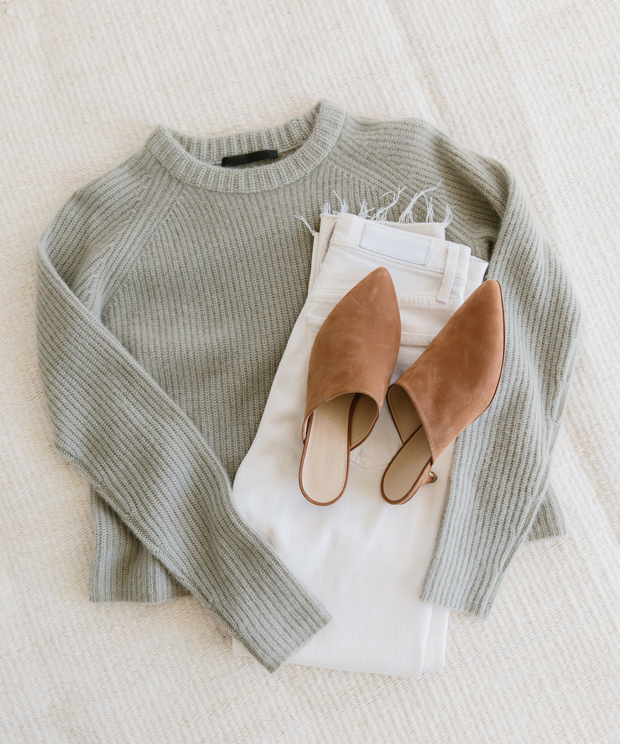A light gray ribbed sweater, white jeans with frayed hems, and stylish brown kitten heel mules are neatly arranged on a cream-colored textured surface.