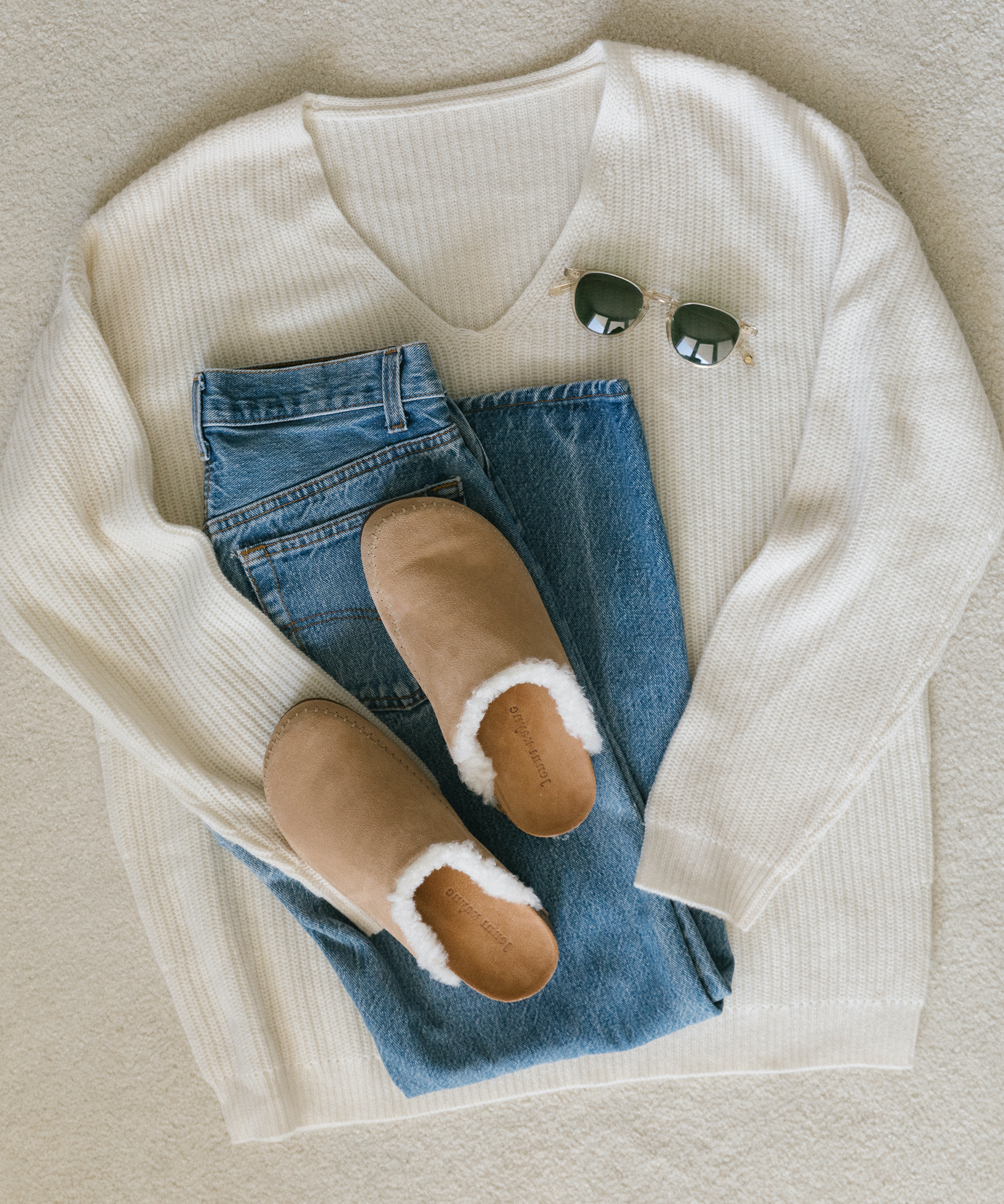 Laydown image of Jenni Kayne cabin sweater, jeans, shearling-lined moc clogs and sunglasses on top of white carpet.