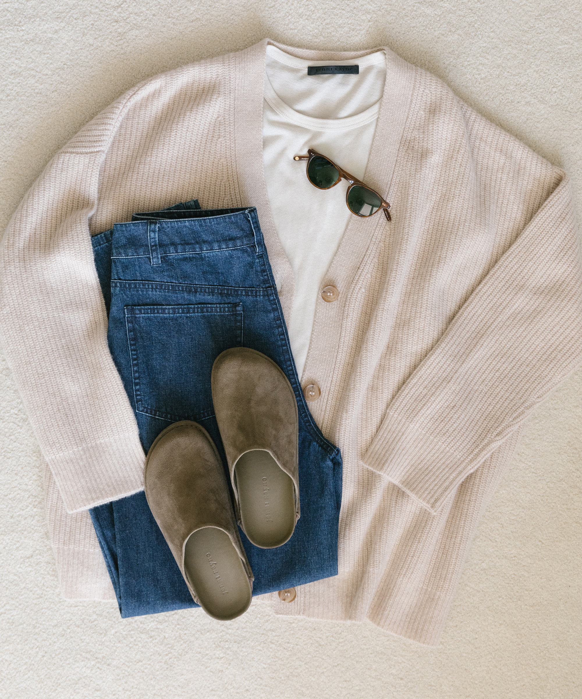 Laydown of Jenni Kayne cashmere cocoon sweater with white tee underneath, denim, moc clogs in suede, and sunglasses on white carpet.