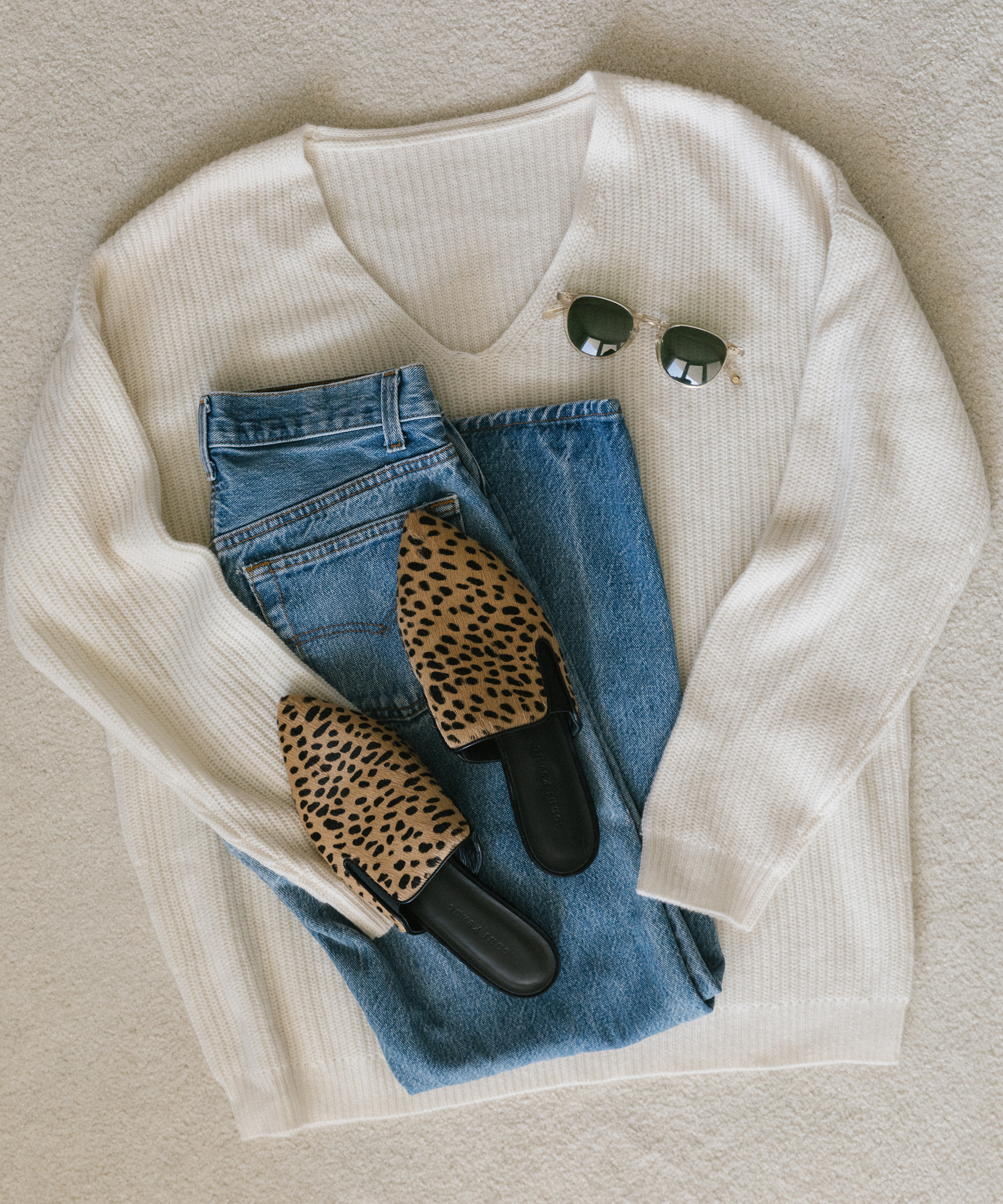 Laydown of Jenni Kayne cabin sweater, cheetah mules, jeans, and sunglasses on white carpet.