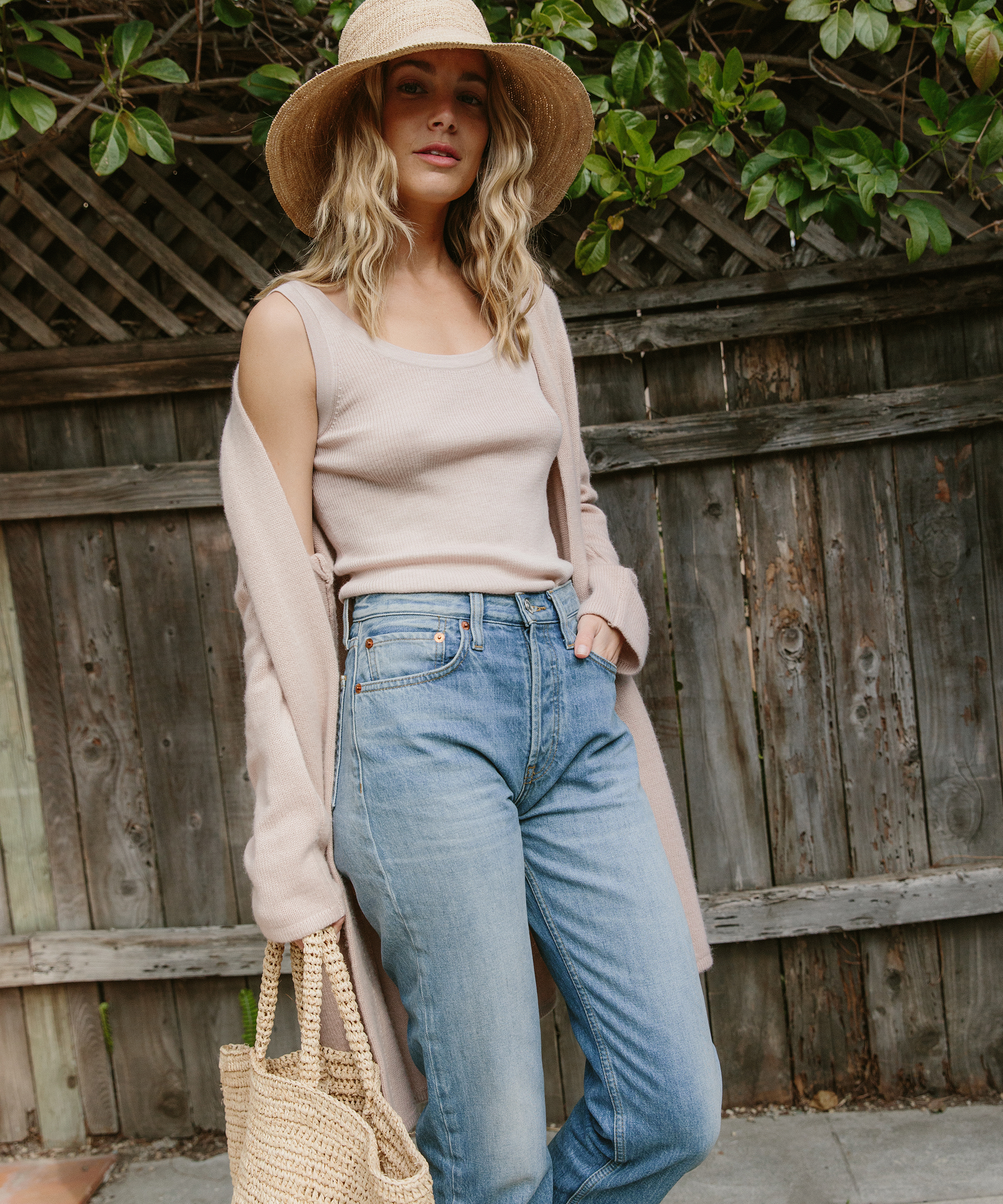Jenni Kayne blonde model wearing blush layering tank, blush cashmere sweater coat, raffia sun hat, and raffia tote bag, and jeans standing outside.