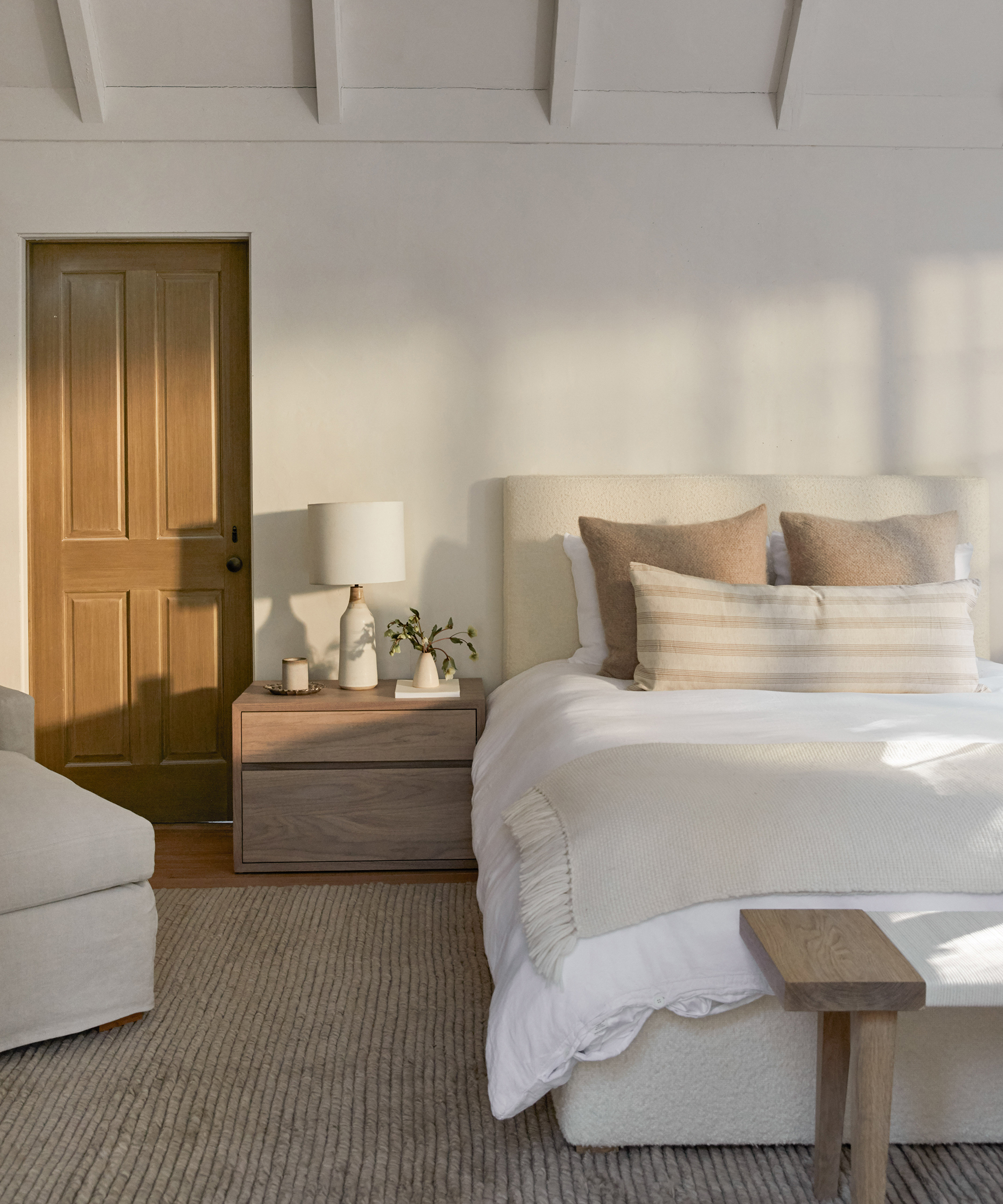 A cozy bedroom with timeless furniture—a beige upholstered bed, layered pillows, a wooden nightstand with a lamp and plant, a soft area rug, and a closed wooden door in the background. Natural light fills the room.