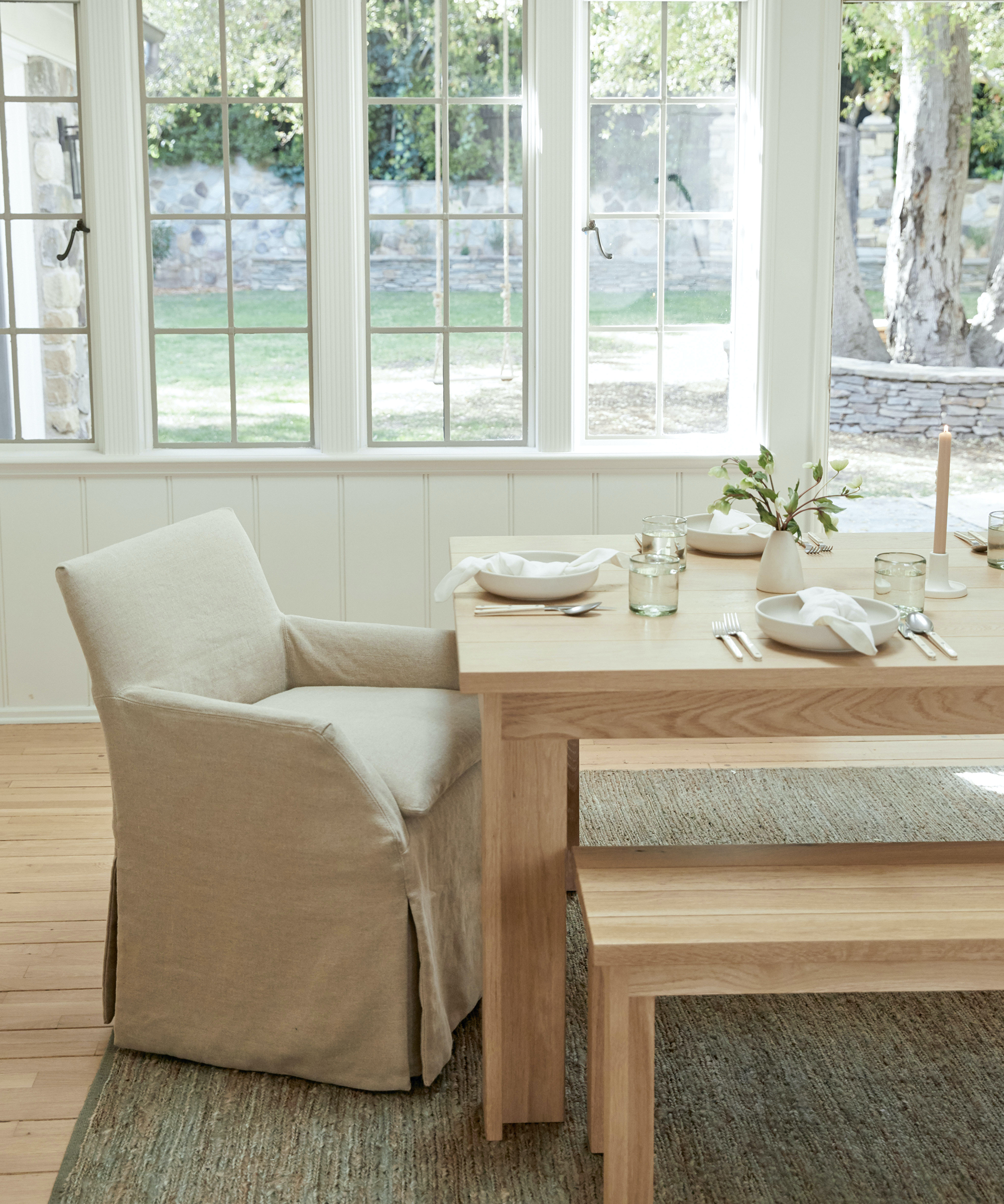 A light-filled dining room with timeless furniture, including a wooden table, matching bench, and slipcovered armchair. The table is set with plates, cutlery, napkins, glasses, and a vase with greenery. Large windows overlook a garden.