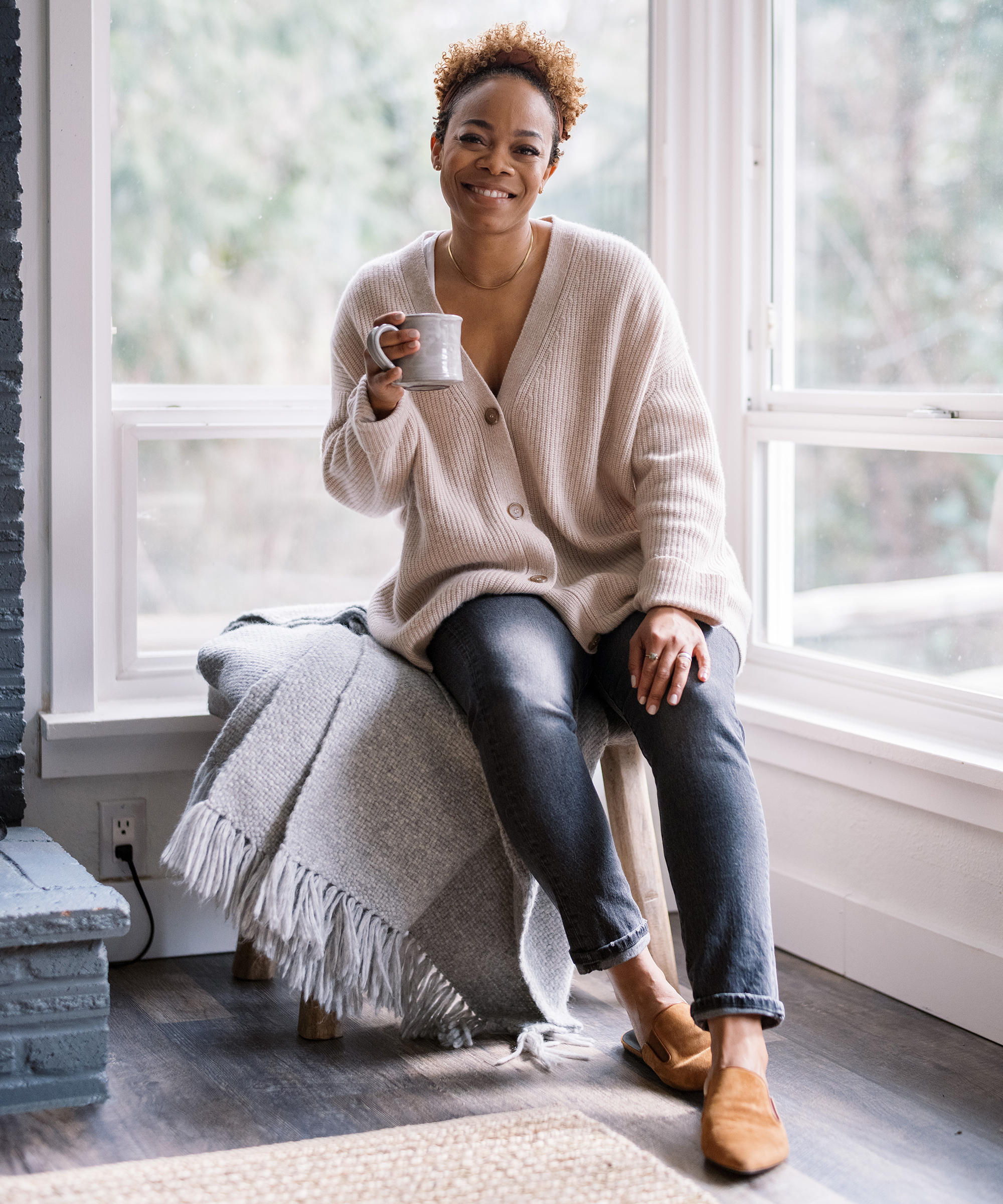 A person with short curly hair sits on a bench by a window in the restoration house, smiling and holding a mug. They wear a cream cardigan, dark jeans, and brown shoes. A gray blanket is draped on the bench beside them.