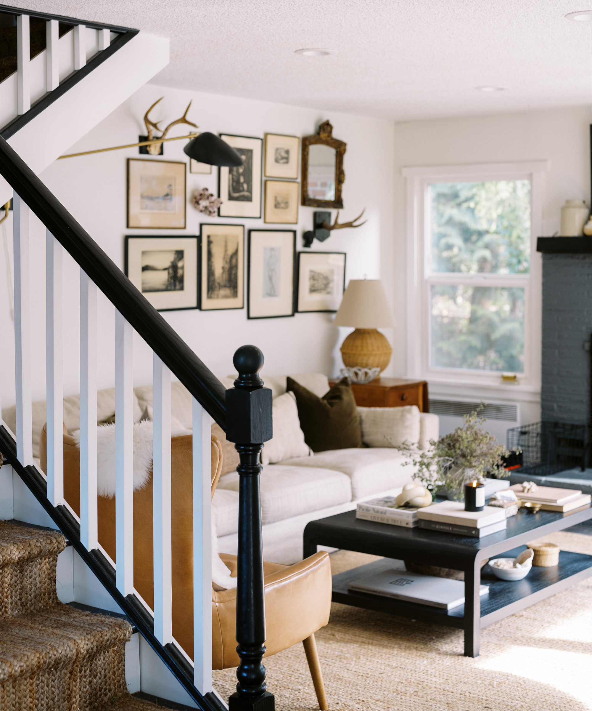 A cozy living room in a restoration house features a beige sofa, brown armchair, black coffee table, gallery wall art, a lamp, and decorative antlers. A staircase with black railing sits in the foreground as light streams through the window.