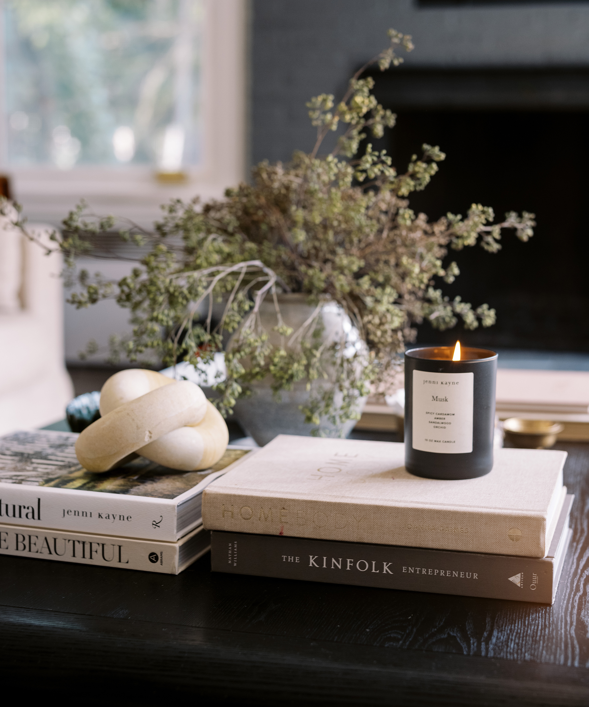 A lit black candle sits on stacked books beside pale decorative stones and a vase with greenery atop a dark coffee table, creating restoration house charm in a cozy, softly lit living room.