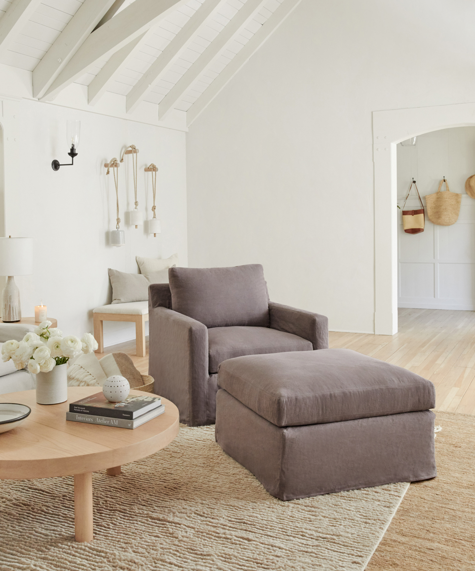 A cozy living room with timeless furniture, including a gray armchair and matching ottoman on a woven rug, a round wooden coffee table with books and flowers, neutral décor, and white walls with wooden beams and natural light.