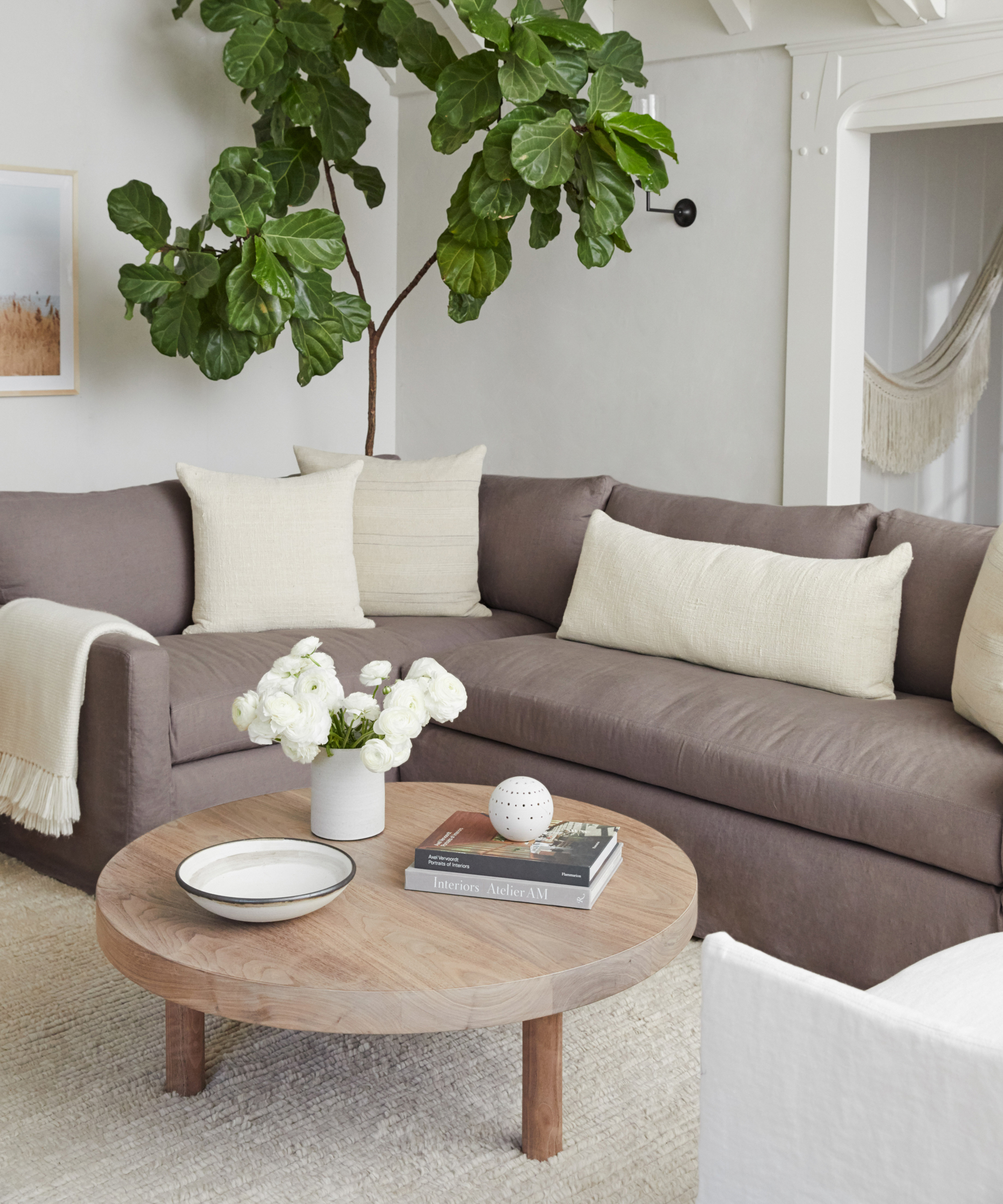 A modern living room featuring timeless furniture, including a large gray sectional sofa, cream pillows, a round wooden coffee table with books and flowers, a potted green plant, and neutral-colored walls and decor.