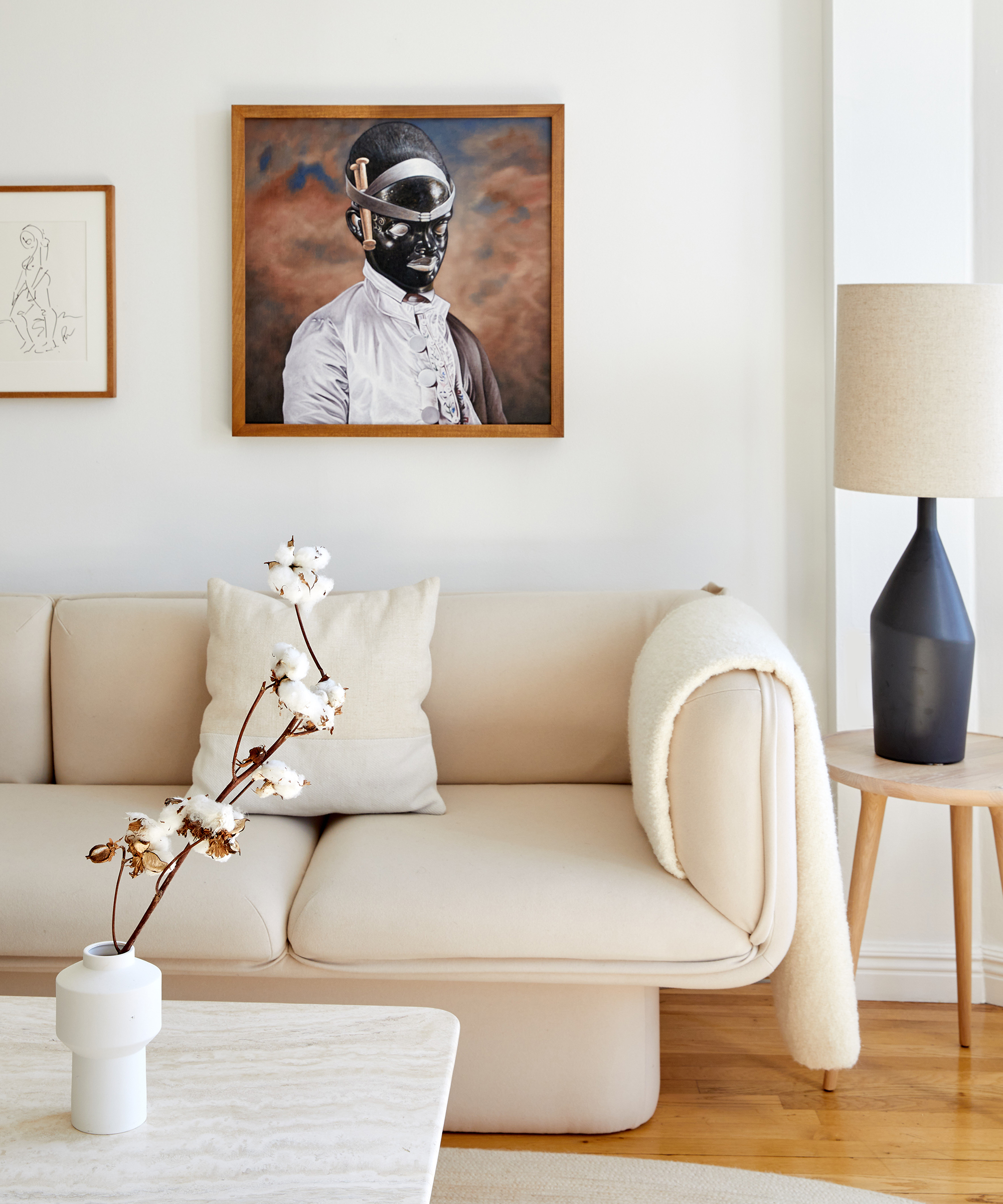 Interior designer Dani Arps' living room with a white sofa with a blanket draped across it, stone coffee table with a vase on top, a side table with a lamp, and art on the walls.