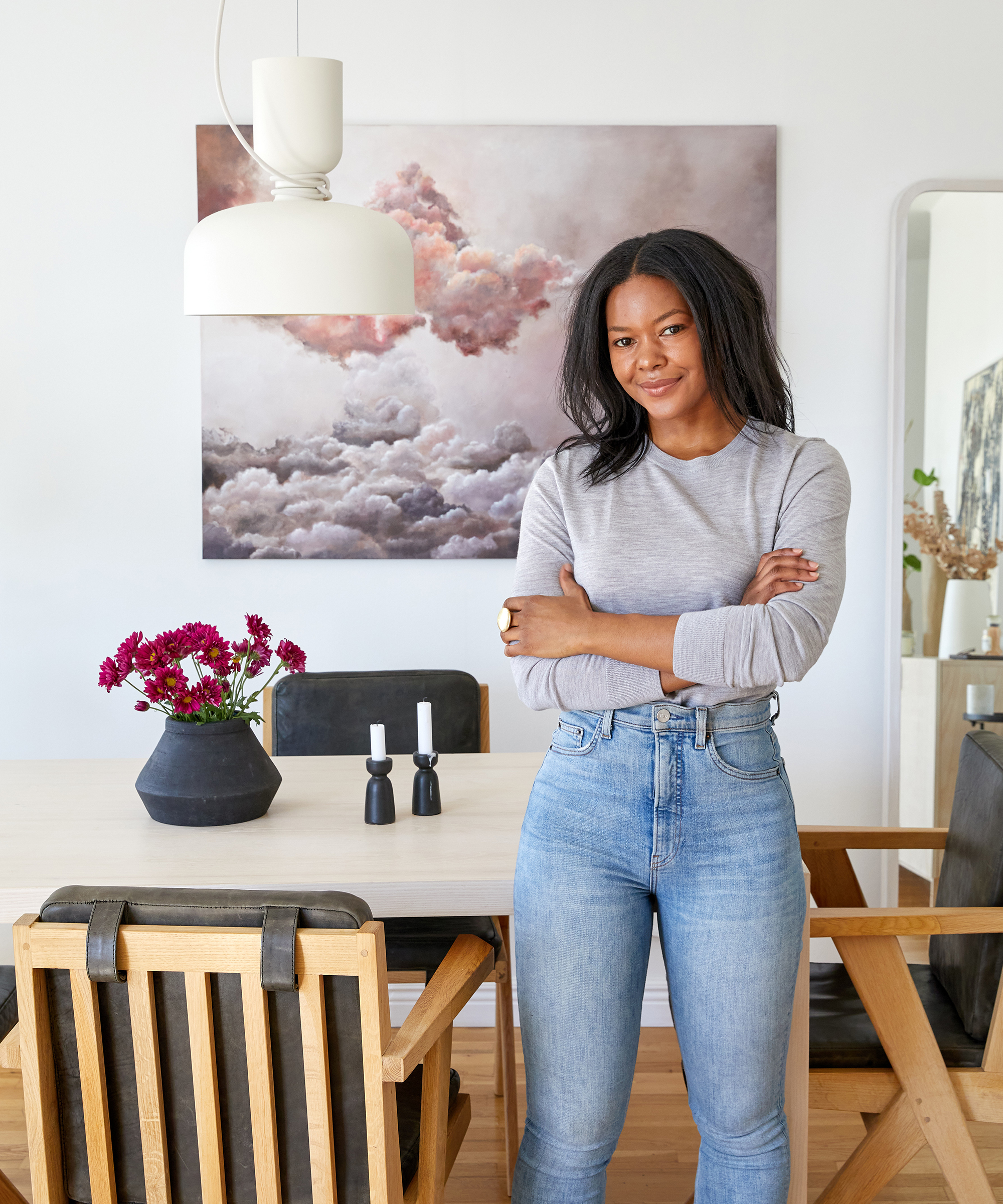 Interior designer Dani Arps standing in her dining room with her arms across her chest in front of a wood table and wood chairs wearing jenni kayne crosby crewneck in heather grey and jeans.