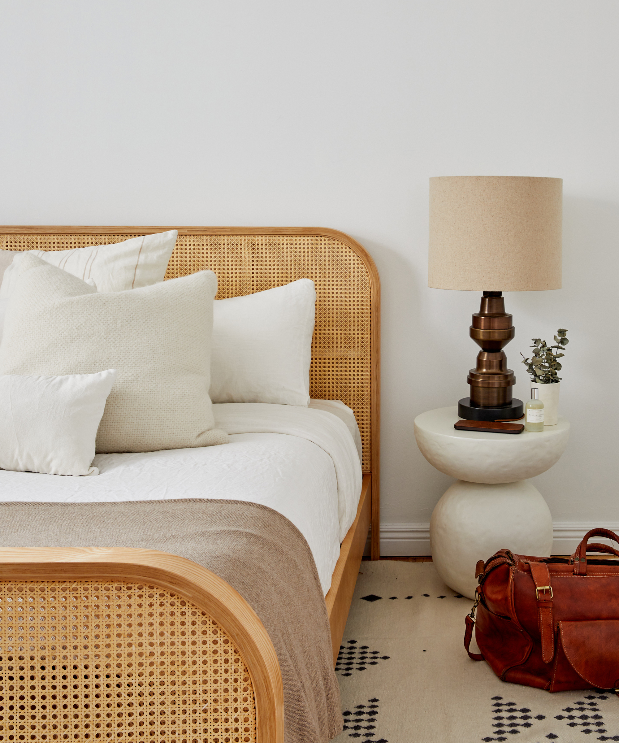Interior designer Dani Arps' bedroom with a wicker bed frame, white linens, jenni kayne alpaca basketweave pillow, a patterned rug, and a stone side table with a lamp on top.