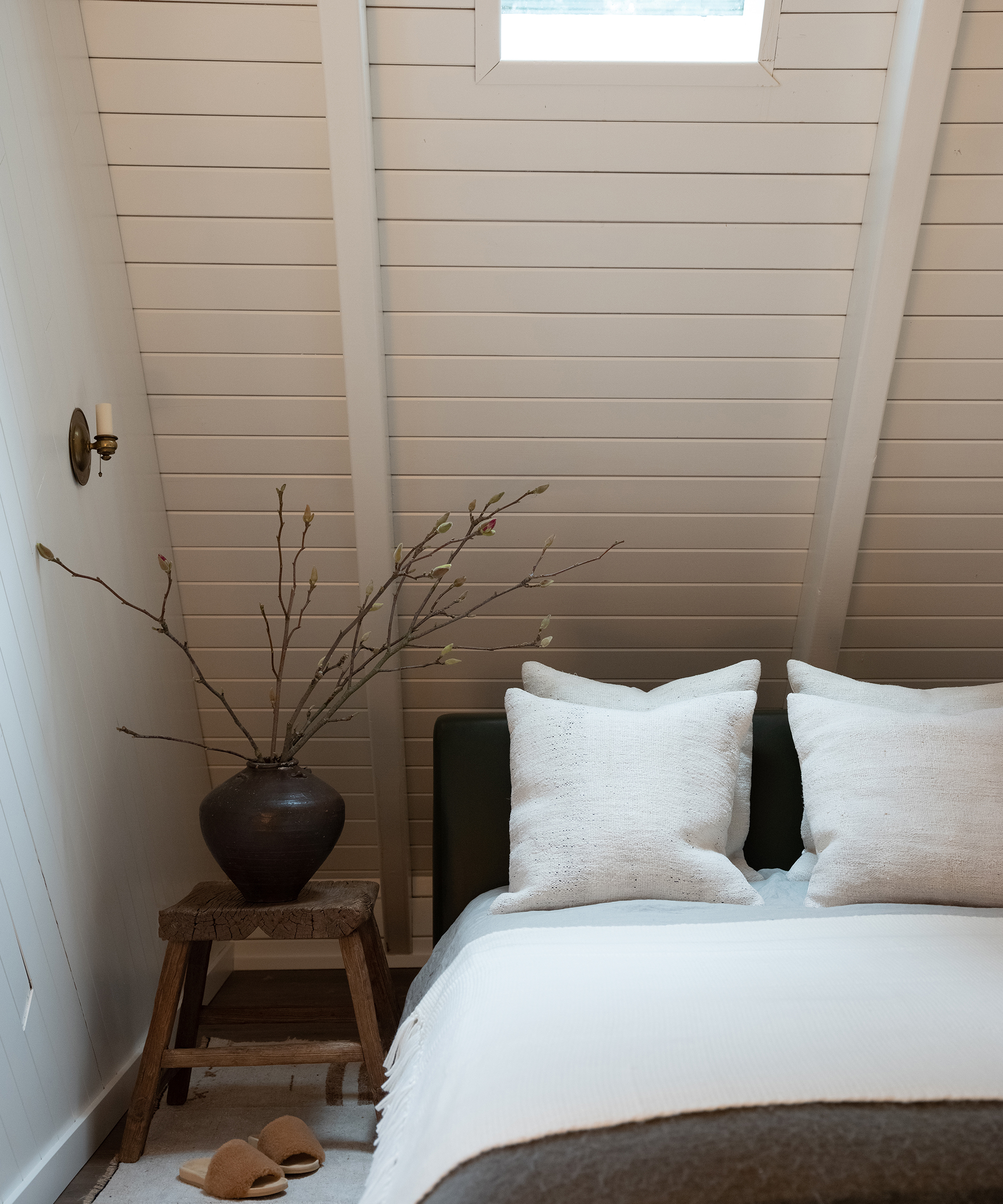 A cozy A-frame cabin bedroom with a slanted white-paneled ceiling, a bed with white pillows and blanket, a wooden stool topped with a dark vase of branches, and slippers resting on the floor near the bed.