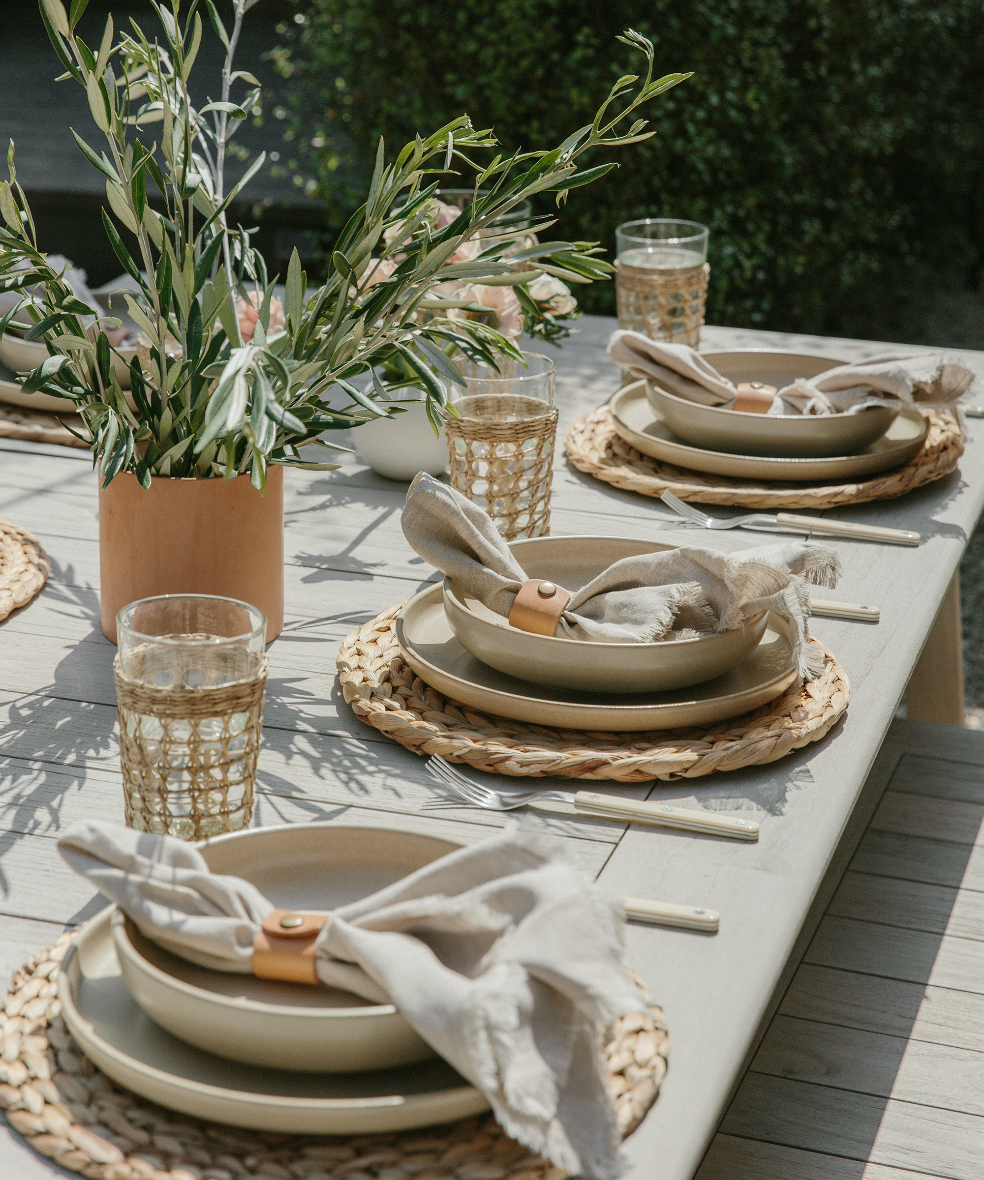 A sunlit outdoor table set for four with neutral-toned plates, beige napkins in leather rings, woven placemats, clear glasses, and a vase of greenery makes for effortless outdoor entertaining ideas.