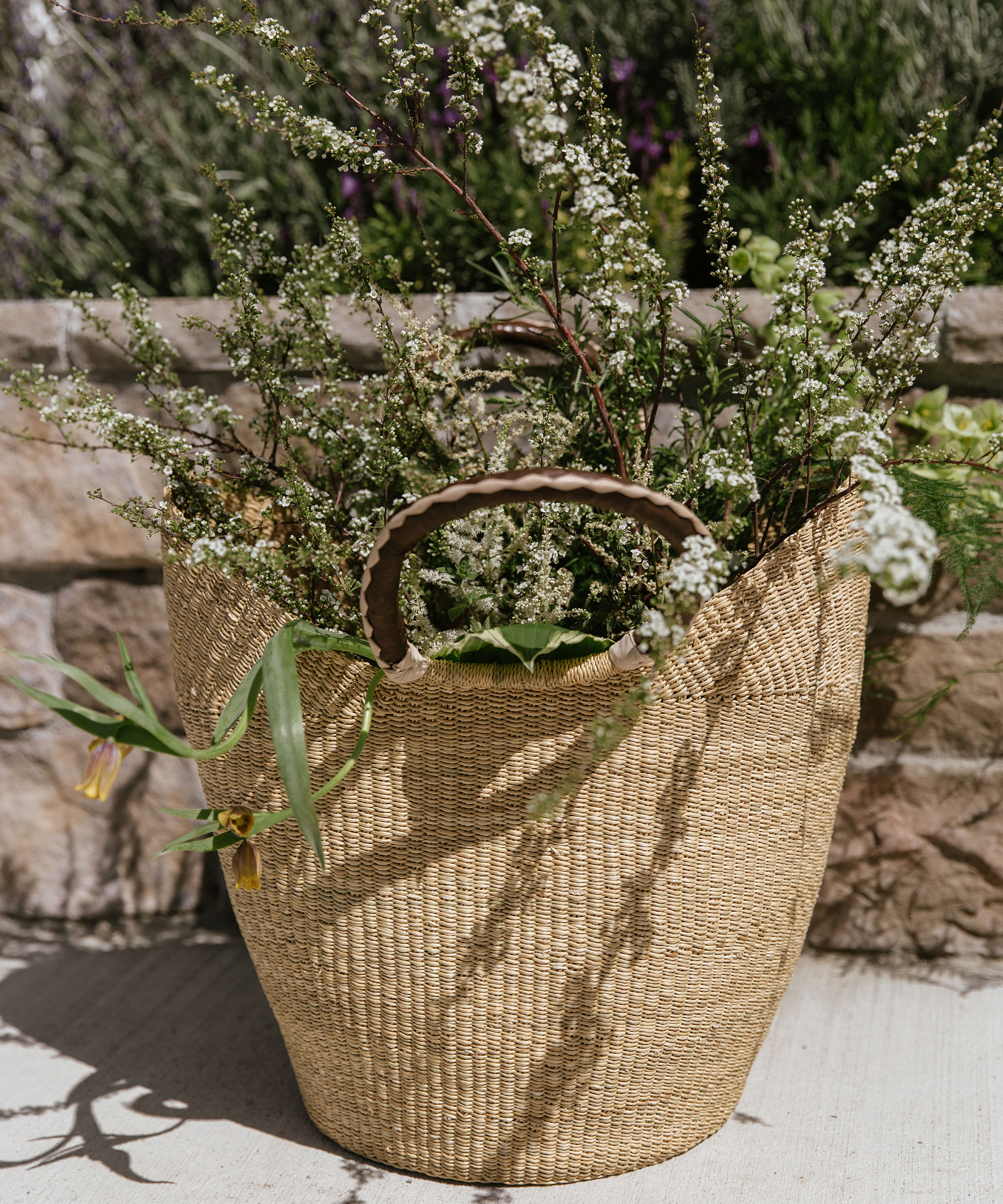A woven straw basket filled with delicate white flowers and green foliage sits on a sunlit stone patio—an inviting touch for outdoor entertaining ideas, framed by a rustic stone wall and lush greenery in the background.