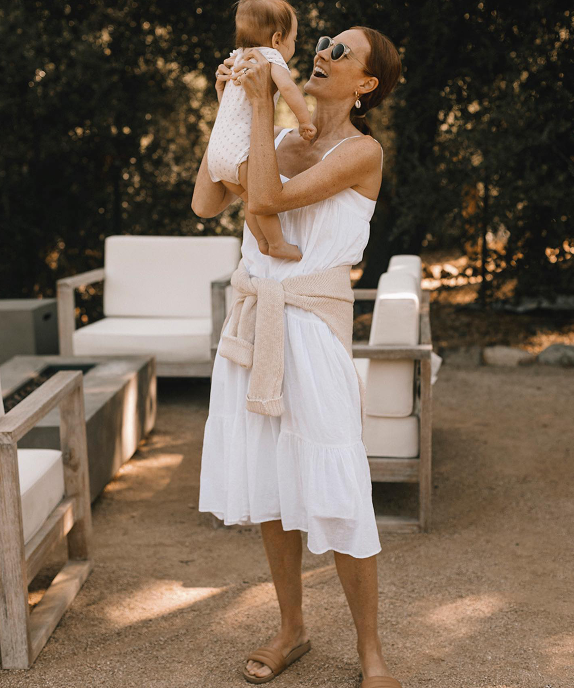 Sam Wennerstrom in the jenni kayne summer dress in white lifting up her daughter and smiling with sunglasses on and a linen sweater tied around her waist.