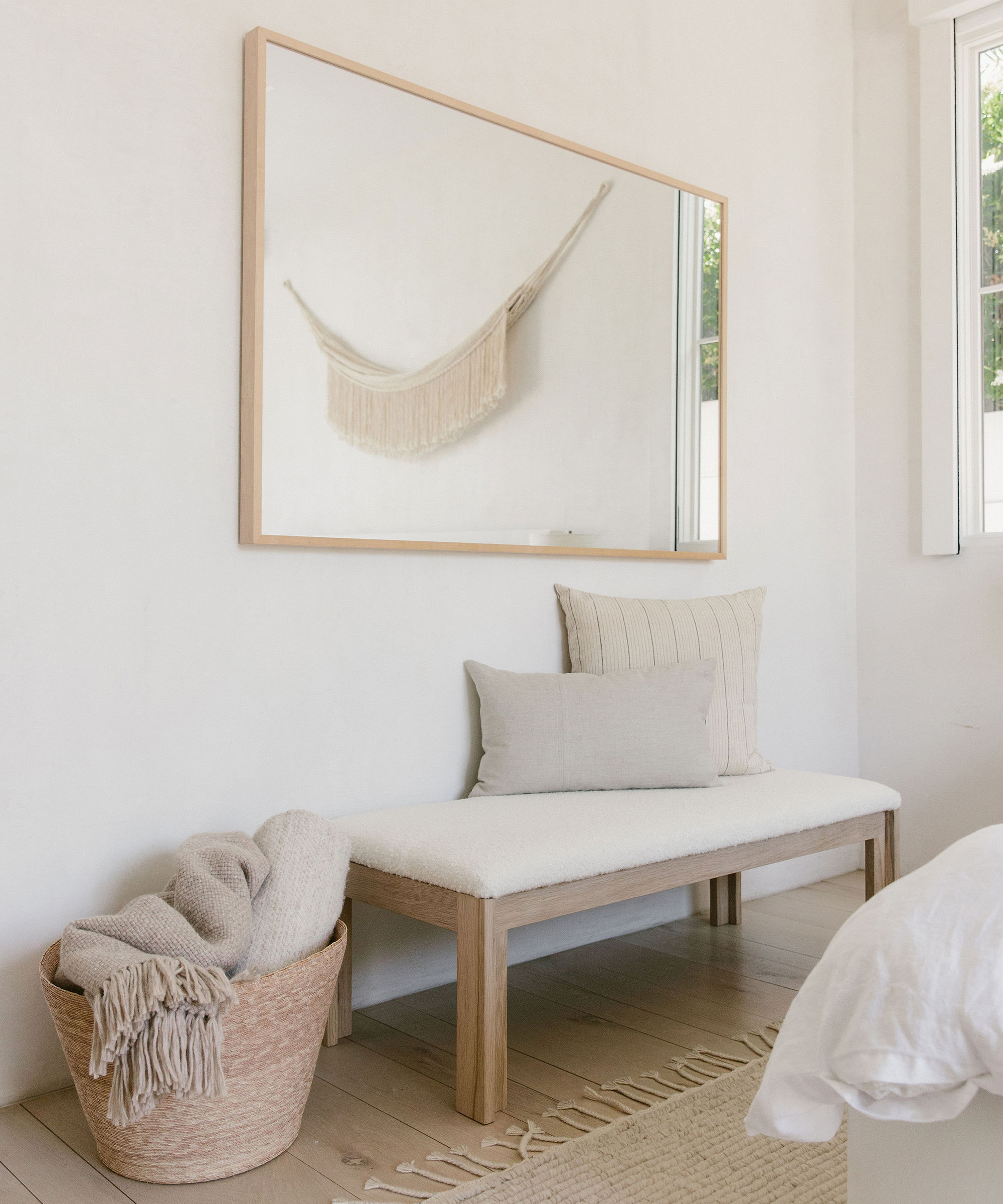 Jenni Kayne upholstered bench in ivory wool boucle underneath a wall mirror with pillows on top and a basket with throws next to it.