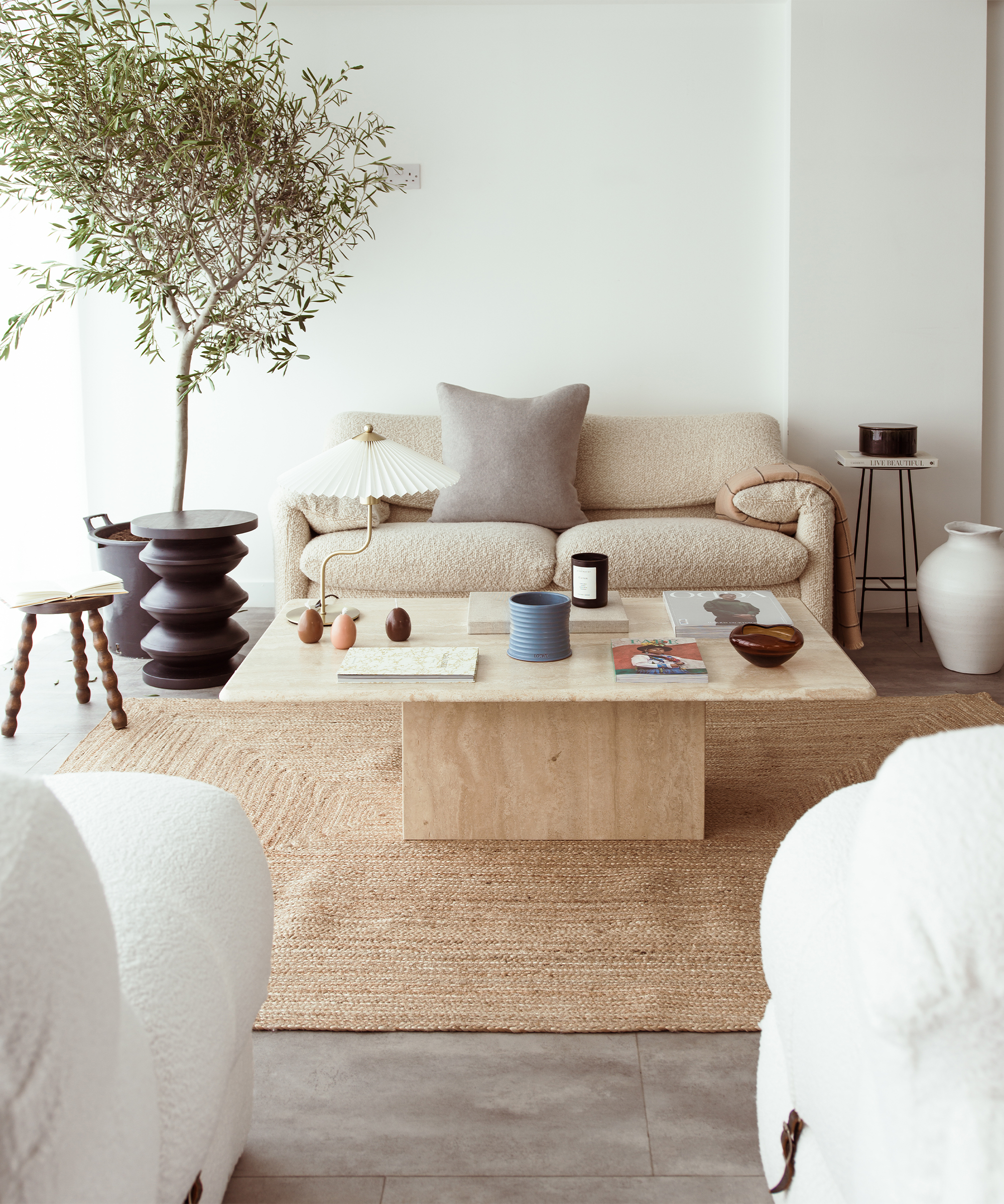 A minimalist living room inspired by Abisola Omole features a beige sofa, neutral pillows, and a light wood coffee table on a woven rug. Decor includes pottery, books, and a potted tree beside the sofa, with soft natural light filling the space.